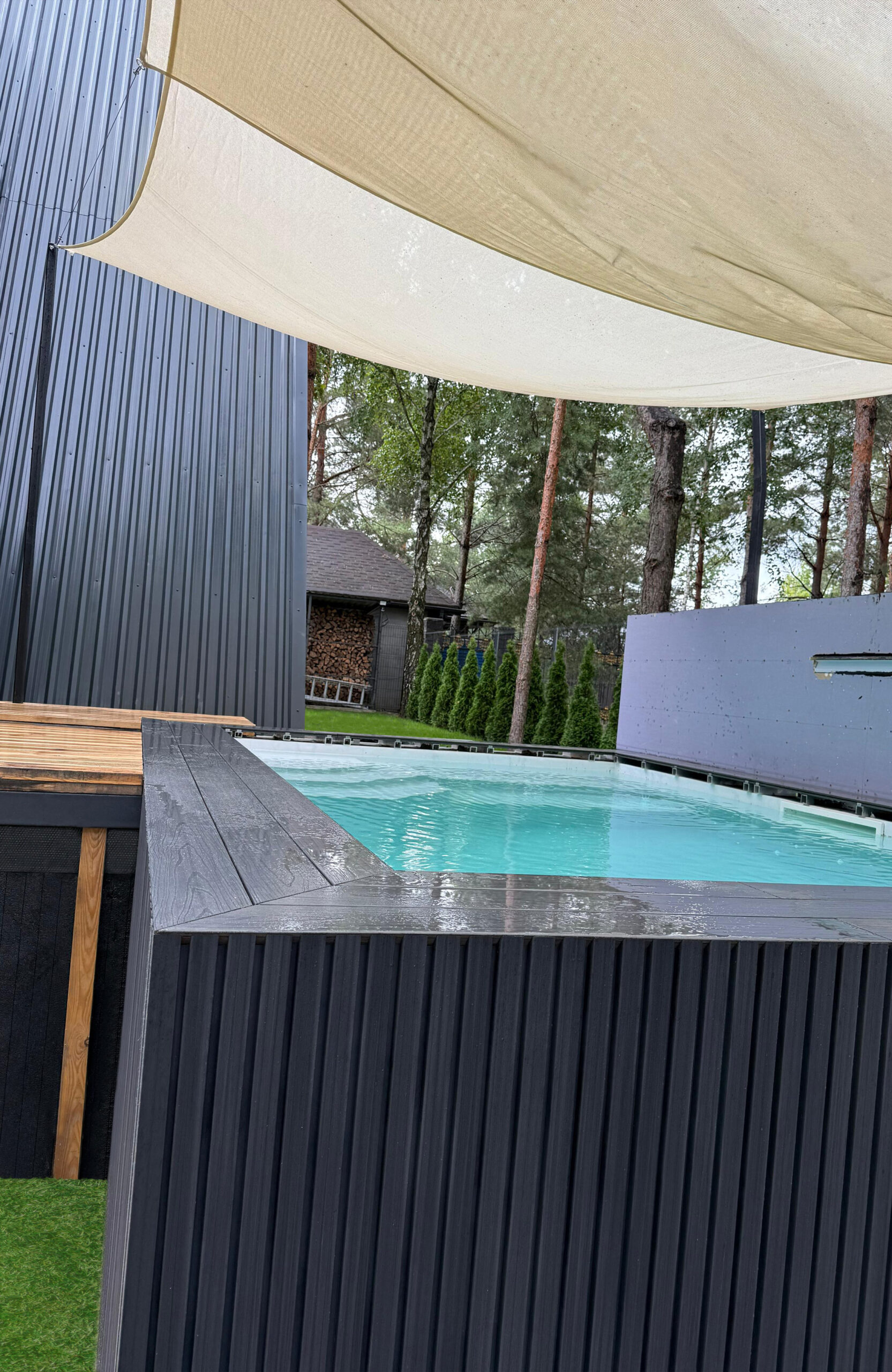 Outdoor pool area with dark wooden walls, a beige shade sail, and tall trees in the background.