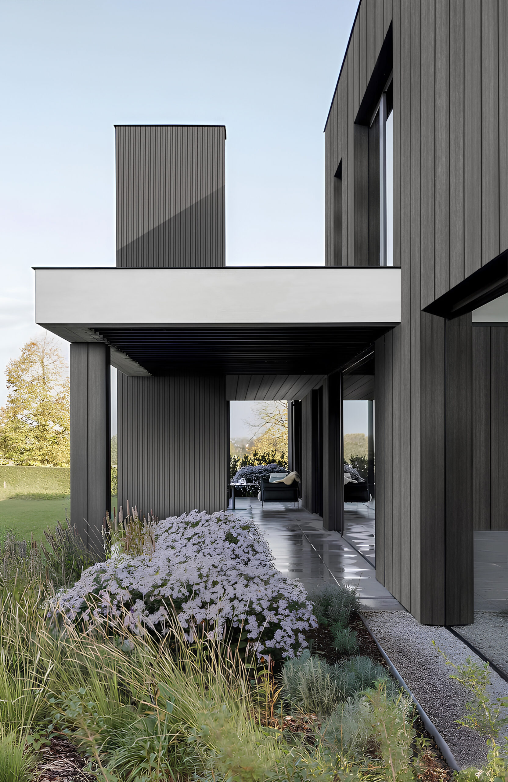 Modern modern house with dark vertical siding, open walkway, and seating area visible through the covered arcades, foregrounded by a garden bed with white flowers