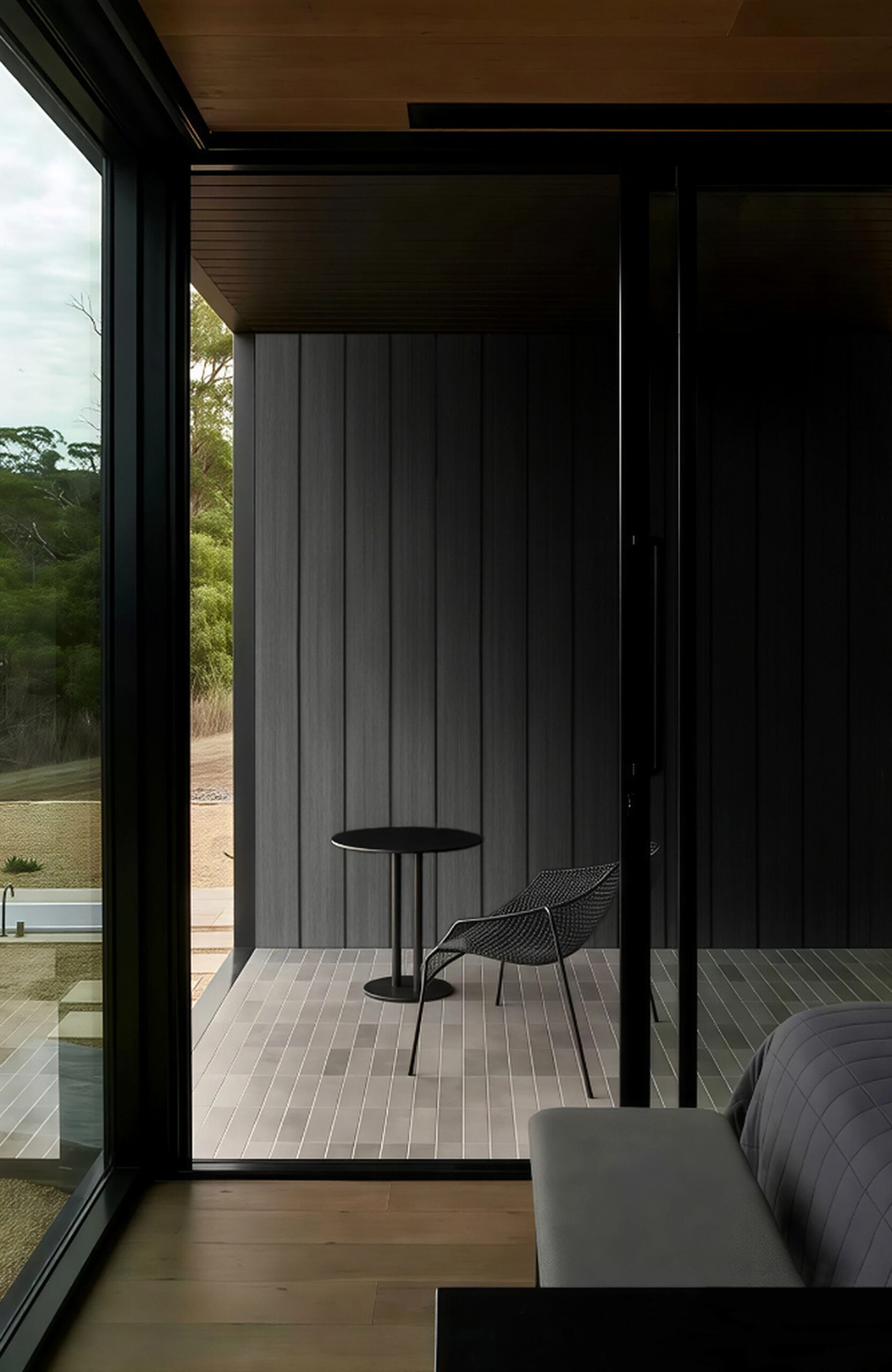Modern indoor balcony seating with a black round table and wire chair by large glass doors, overlooking greenery outside.