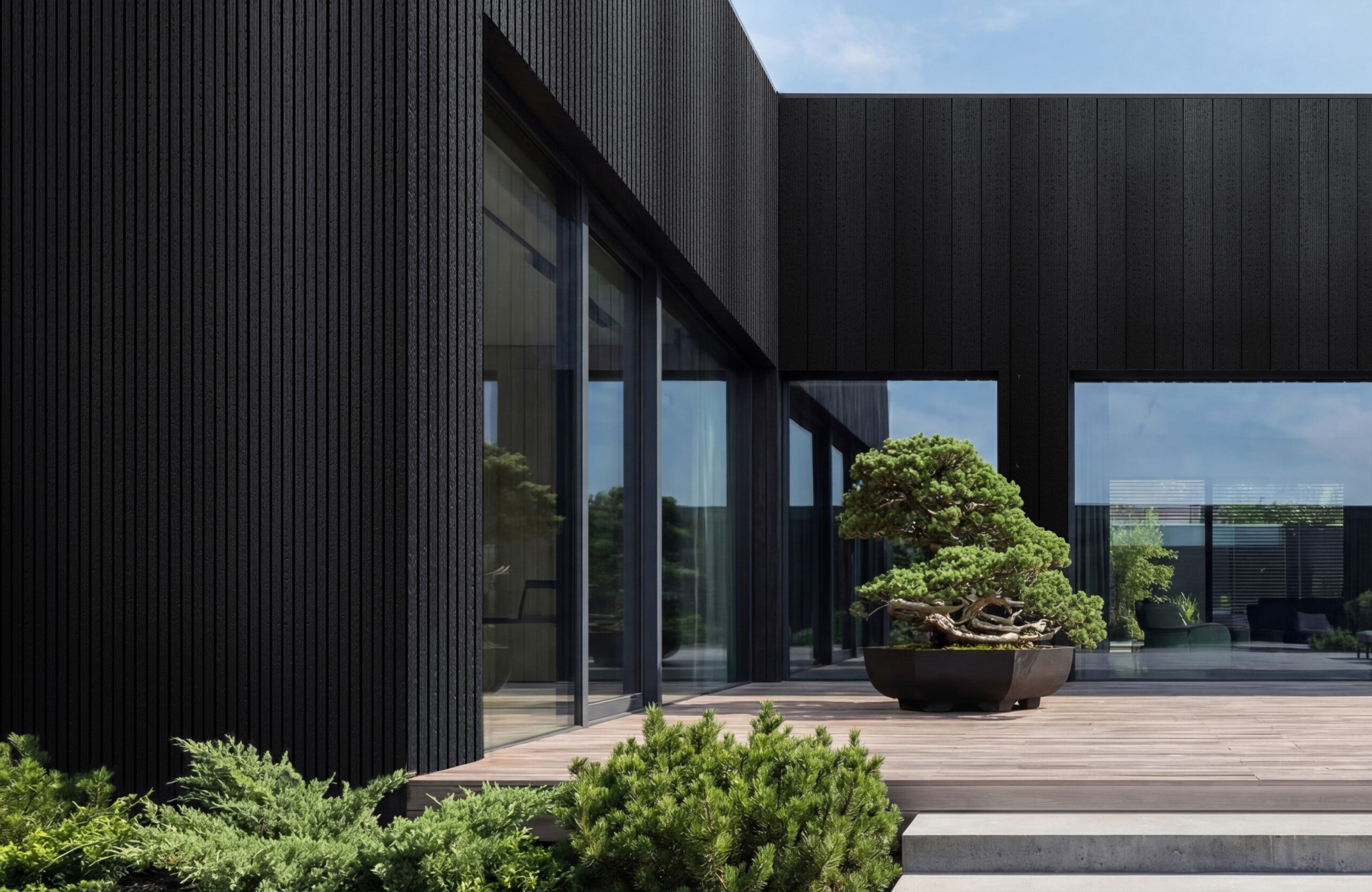 Modern black-clad building with tall glass windows and a wooden deck, overlooking neatly trimmed greenery and a potted bonsai tree.