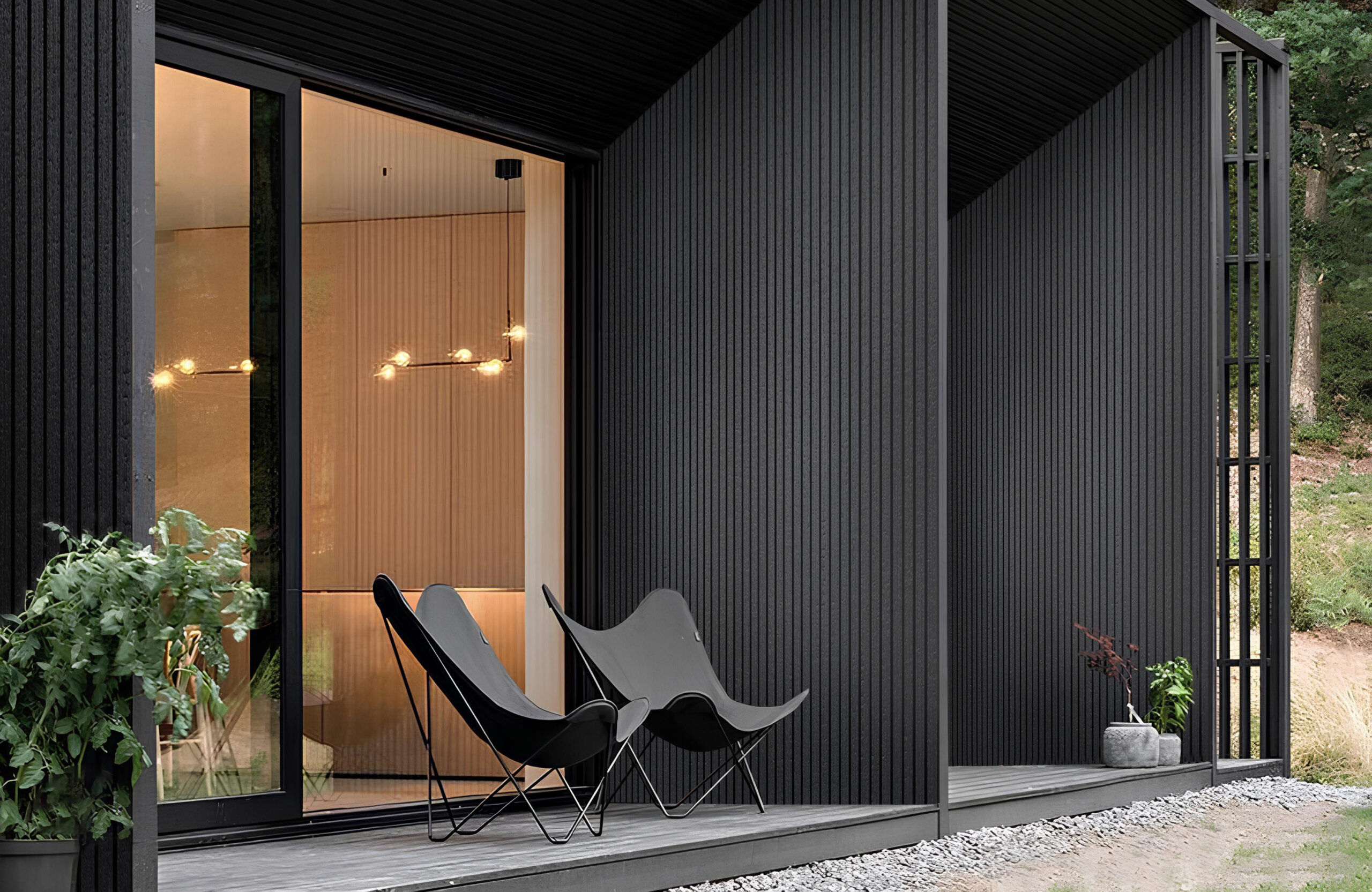 Modern dark wood exterior with glass sliding doors and a warmly lit interior; black mid-century chairs on a wooden deck. Outside: greenery and gravel path nearby.