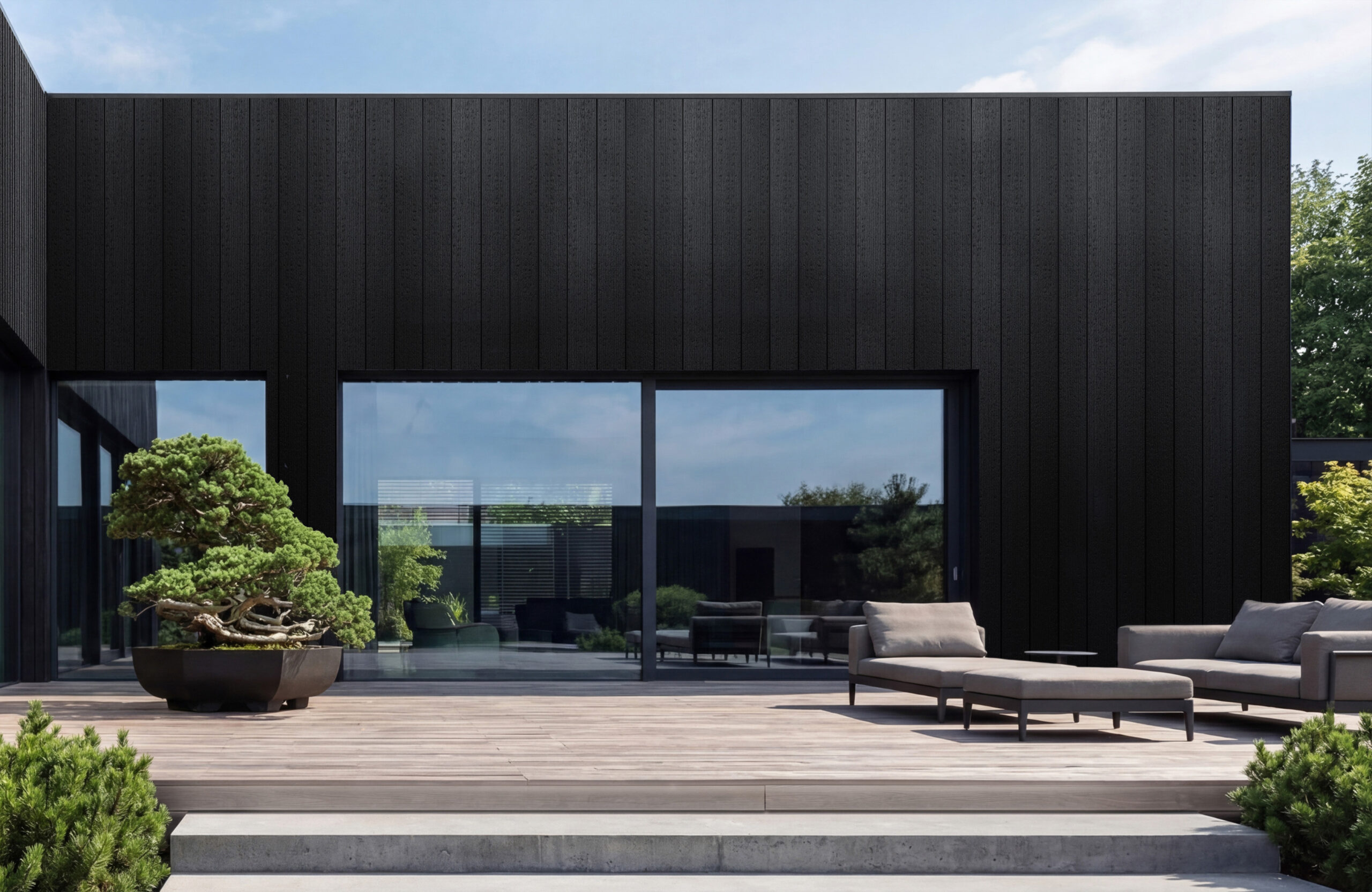 Modern black-clad building with large glass doors and an outdoor lounge on a wooden deck, a bonsai-style tree in the foreground.