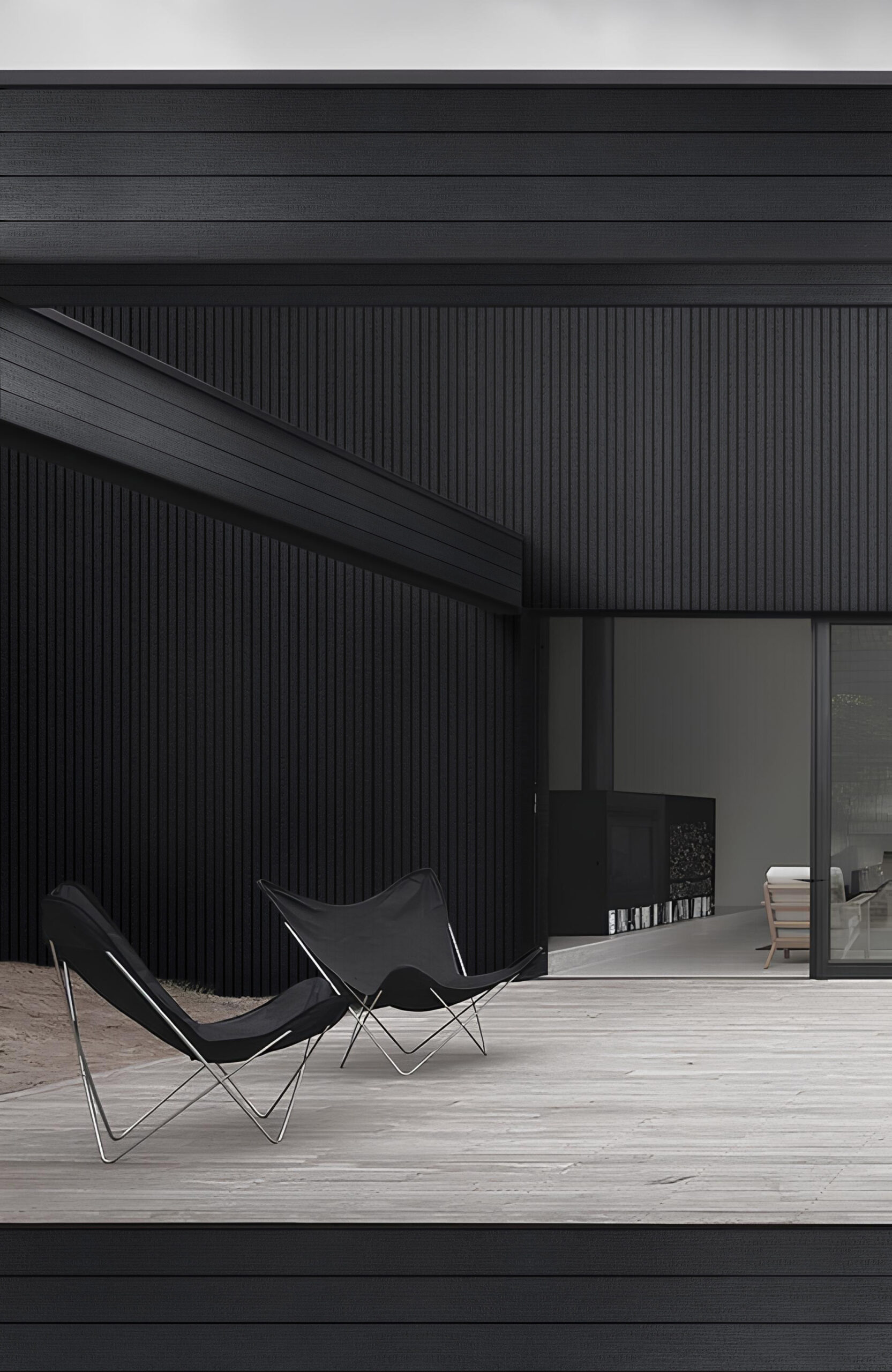 Modern dark-clad house exterior with a light concrete patio and black lounge chairs facing a glass door/indoor seating area