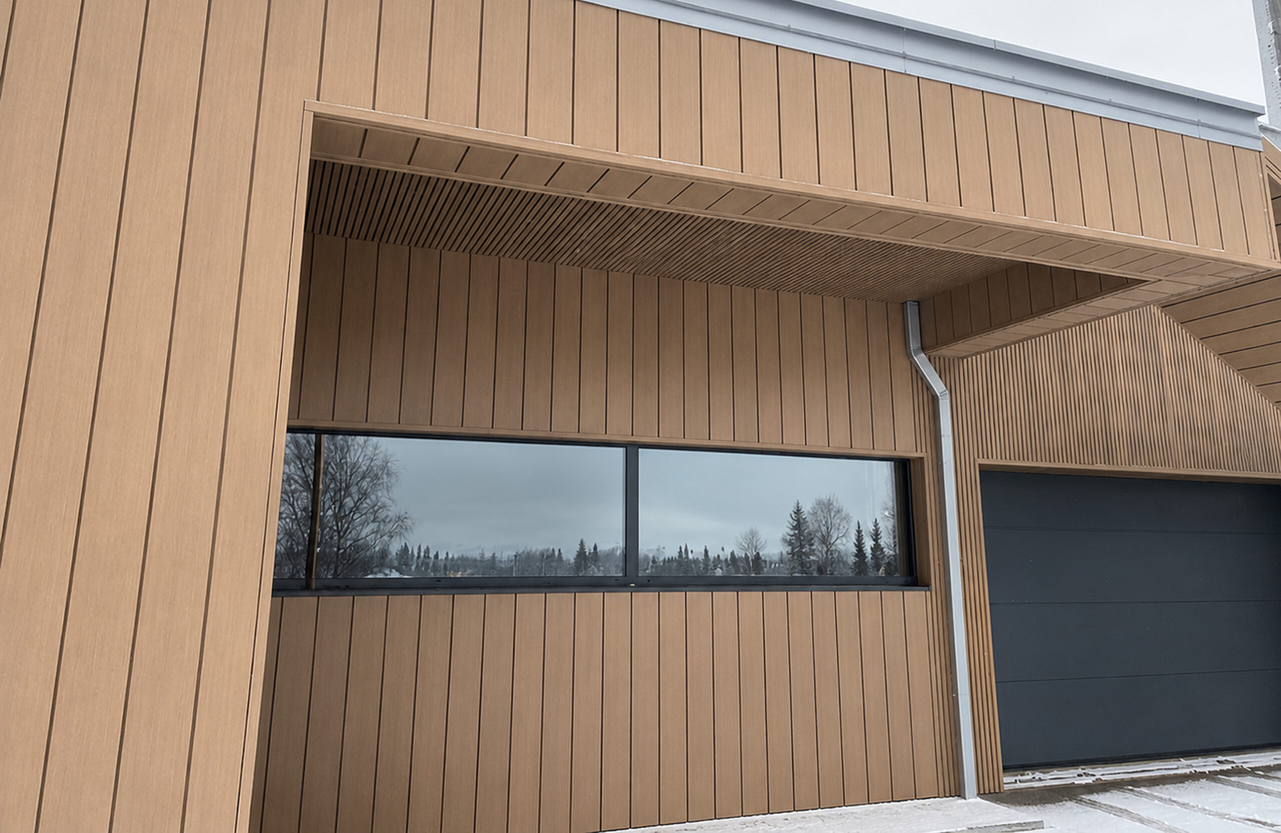 Wood-clad building facade with a long horizontal window and metal gutter, snow on the ground outside.