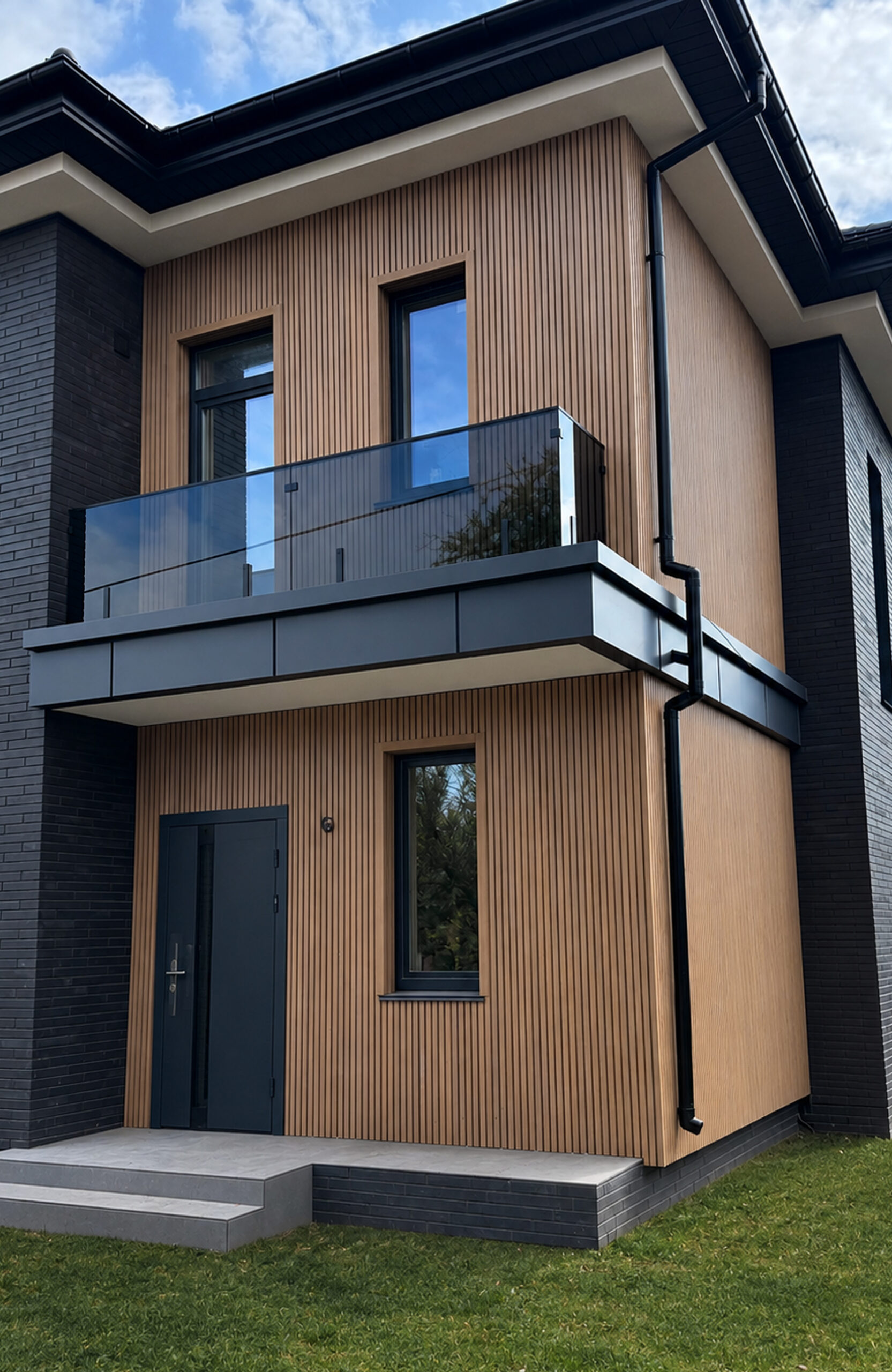 Modern two-story home with vertical wooden siding, dark brick accents, and a glass balcony railing on the upper level.