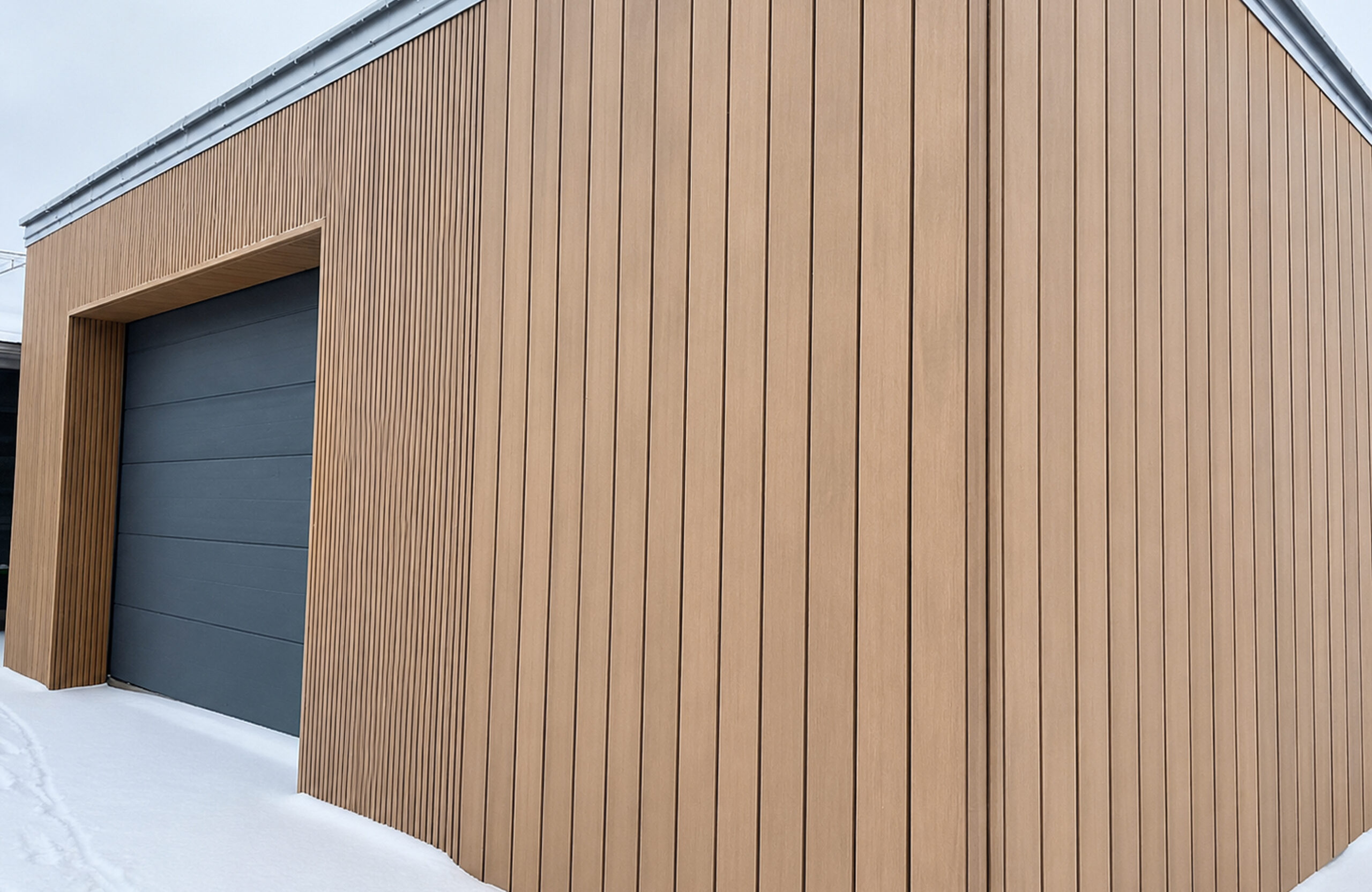 Brown vertical wood siding covers the wall beside a dark blue garage door; snow on the ground nearby.