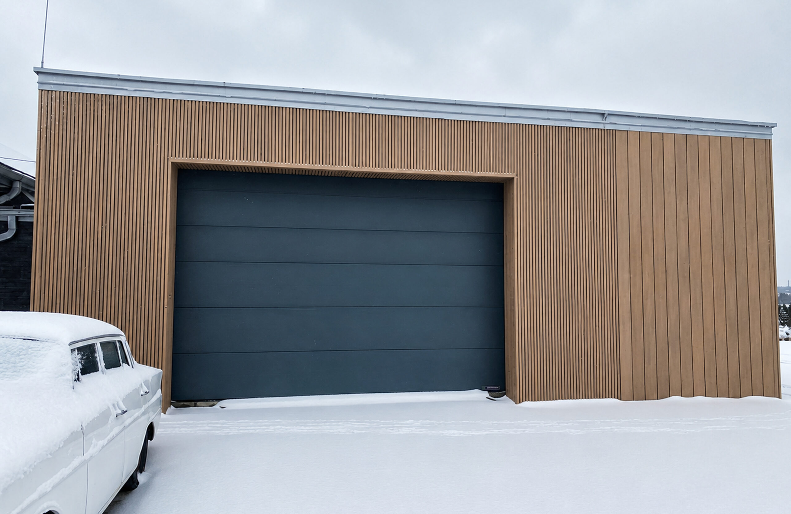 Snowy exterior of a wooden-clad garage with a large dark blue door; a snow-covered car is parked in the foreground.