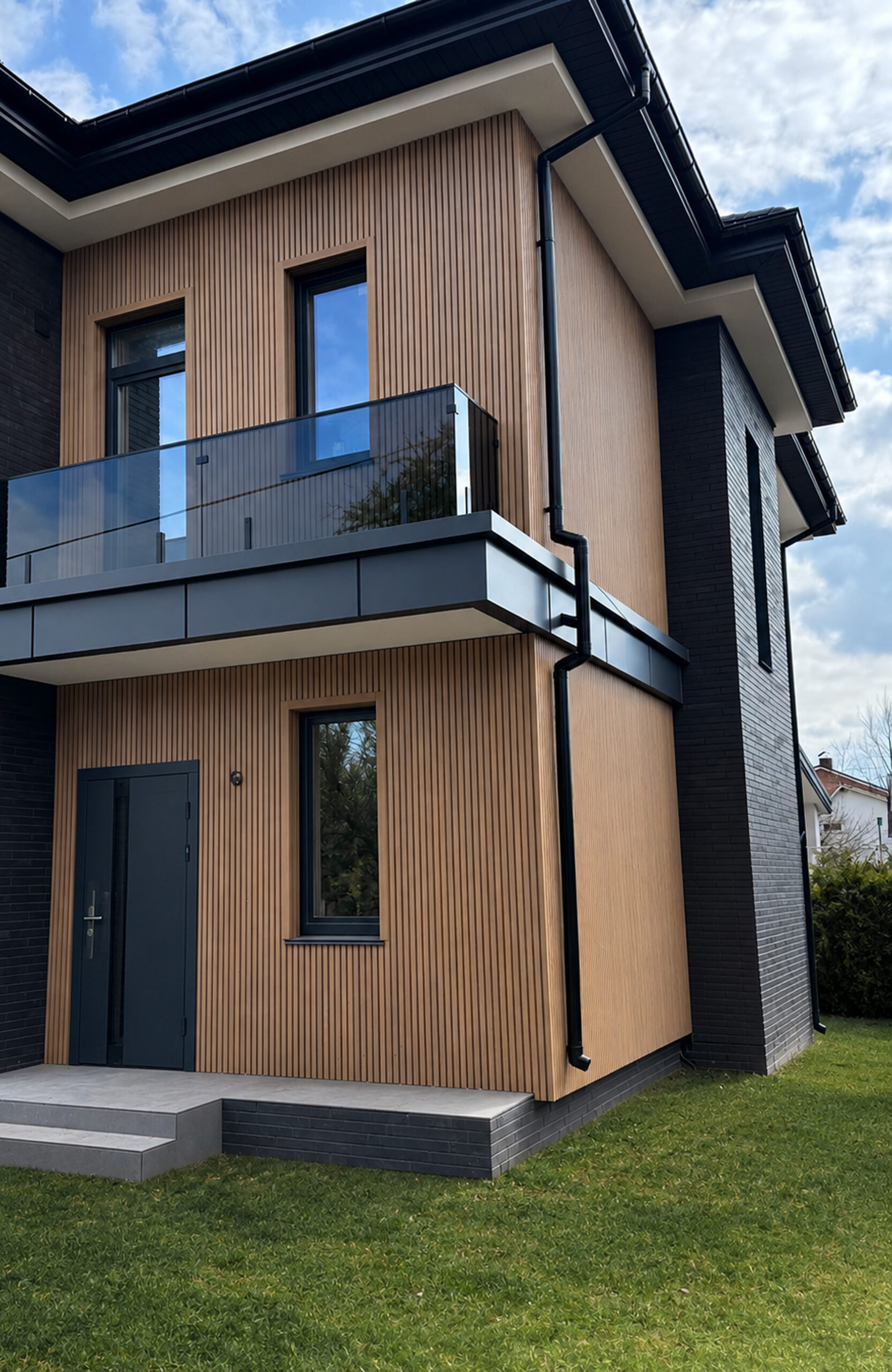 Contemporary two-story house with vertical wood siding, dark window frames, and glass balcony railing.
