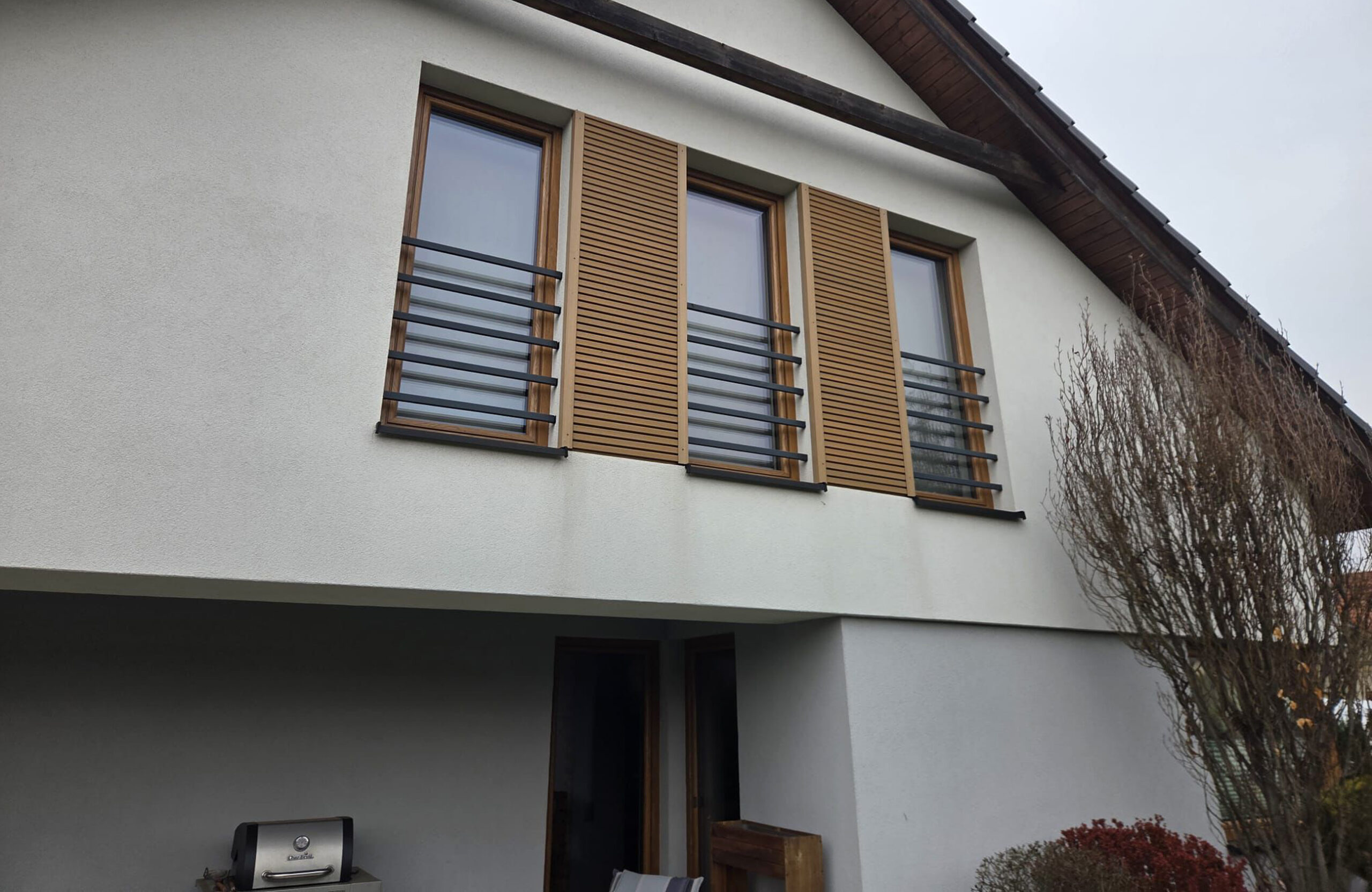 Facade of a modern house with two large windows and wooden shutters, and black metal balcony rails along the wall.