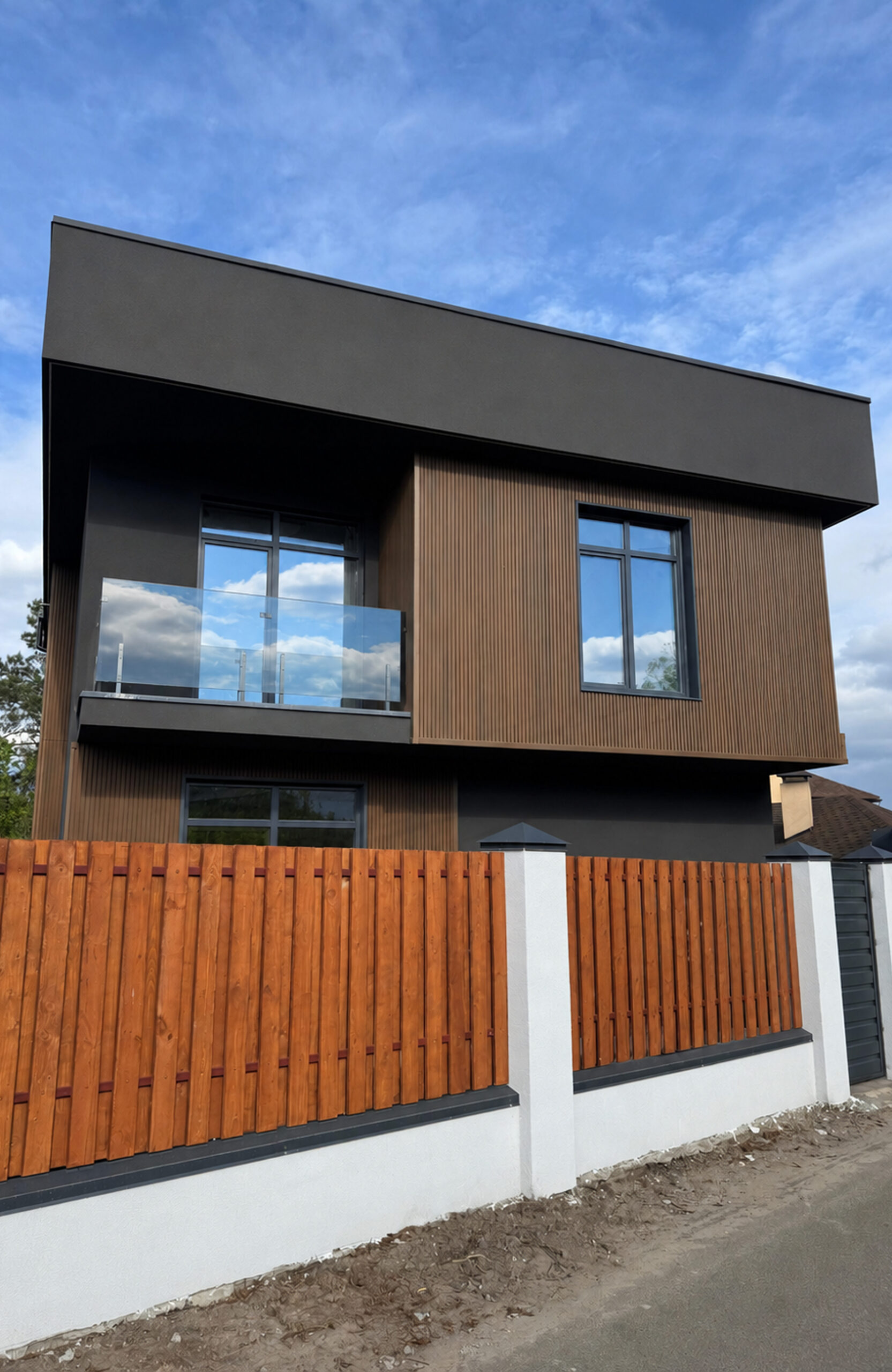 Modern two-story house with dark gray upper volume, brown vertical wood siding, and a glass balcony railing against a blue sky.
