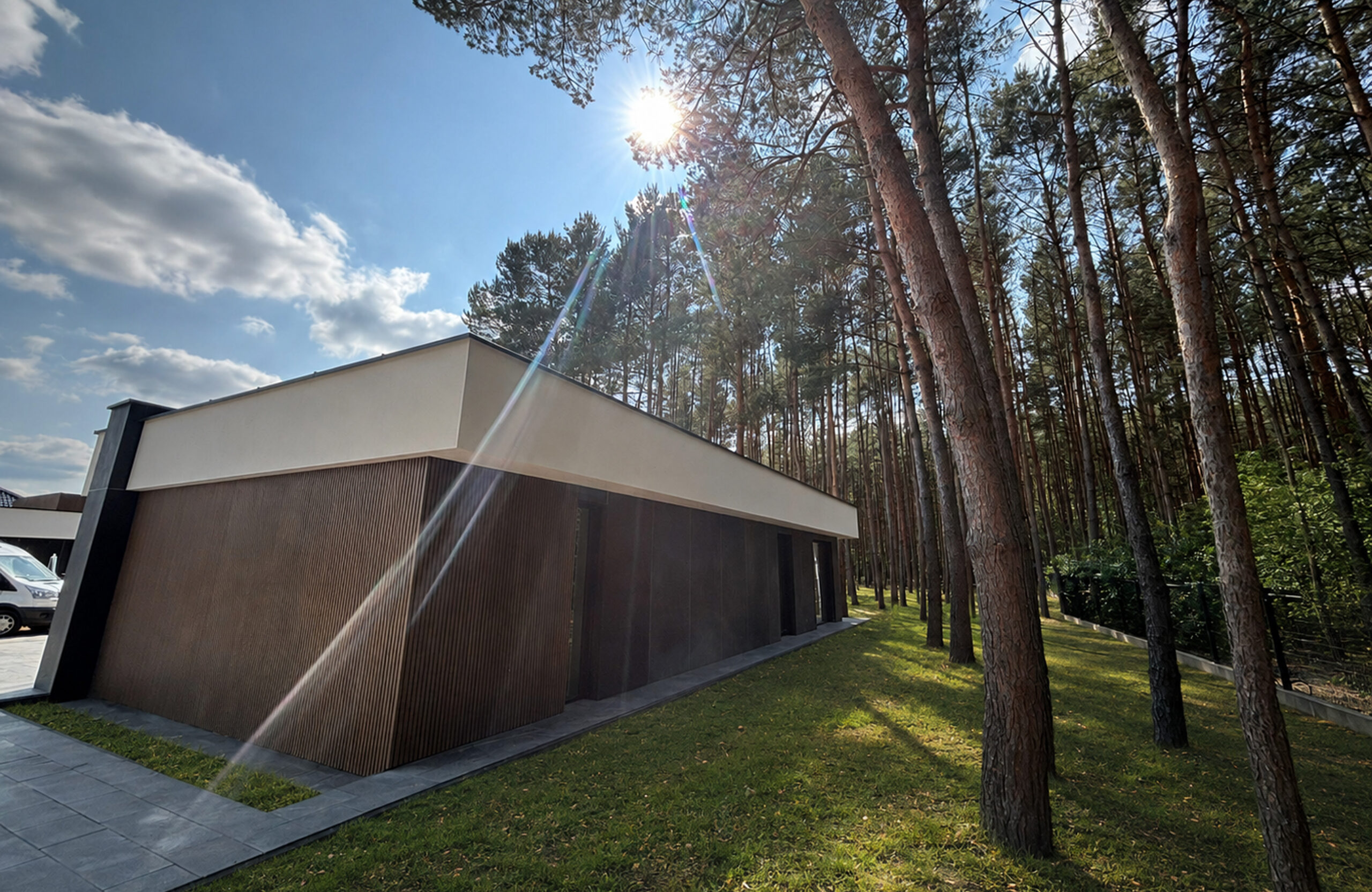 Modern building with dark brown siding beside a dense pine forest under a bright sun.