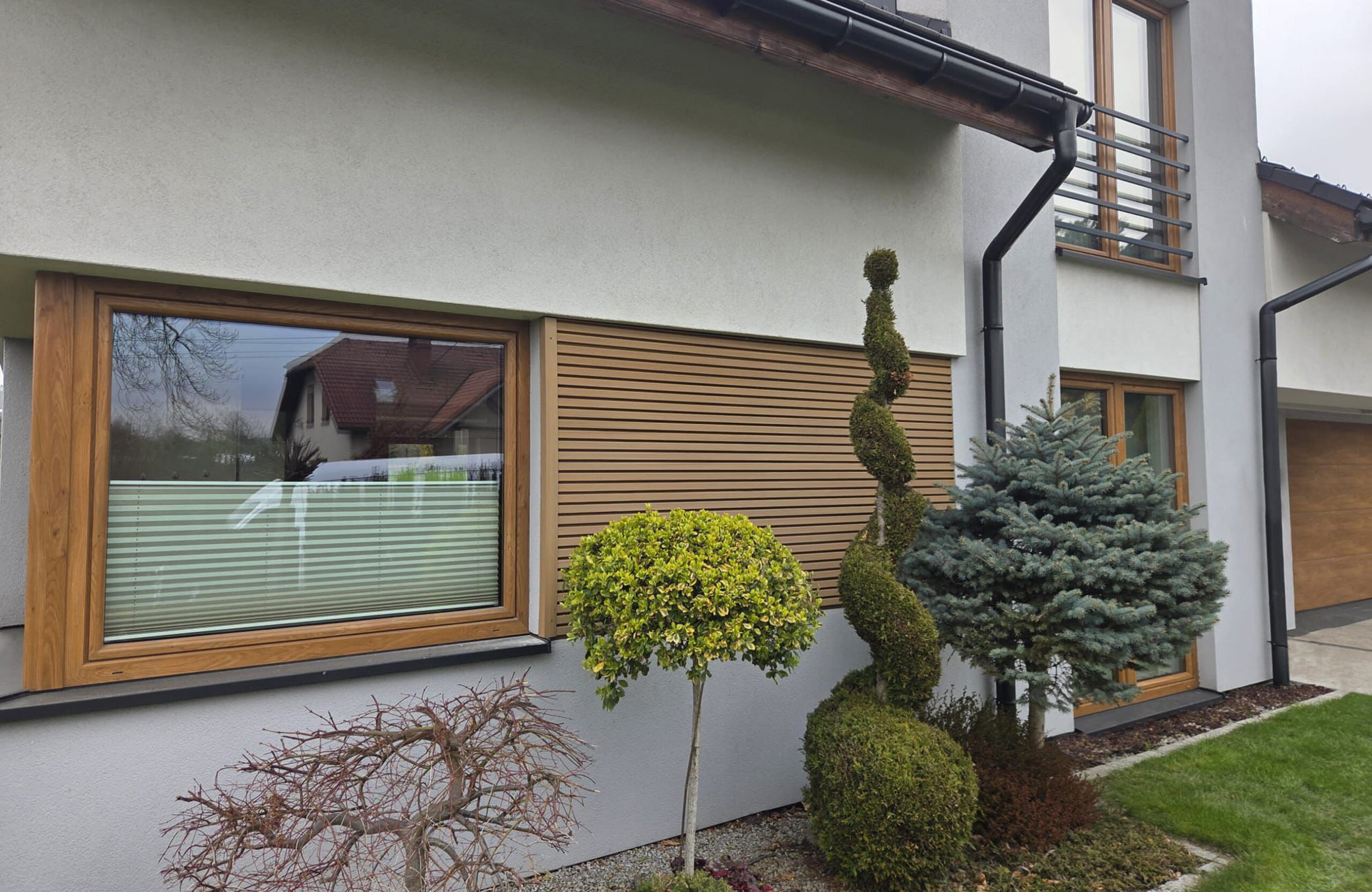 Modern house facade with wooden-framed window, horizontal wooden shutters, and gray stucco walls surrounded by trimmed spiraled topiary and evergreens.