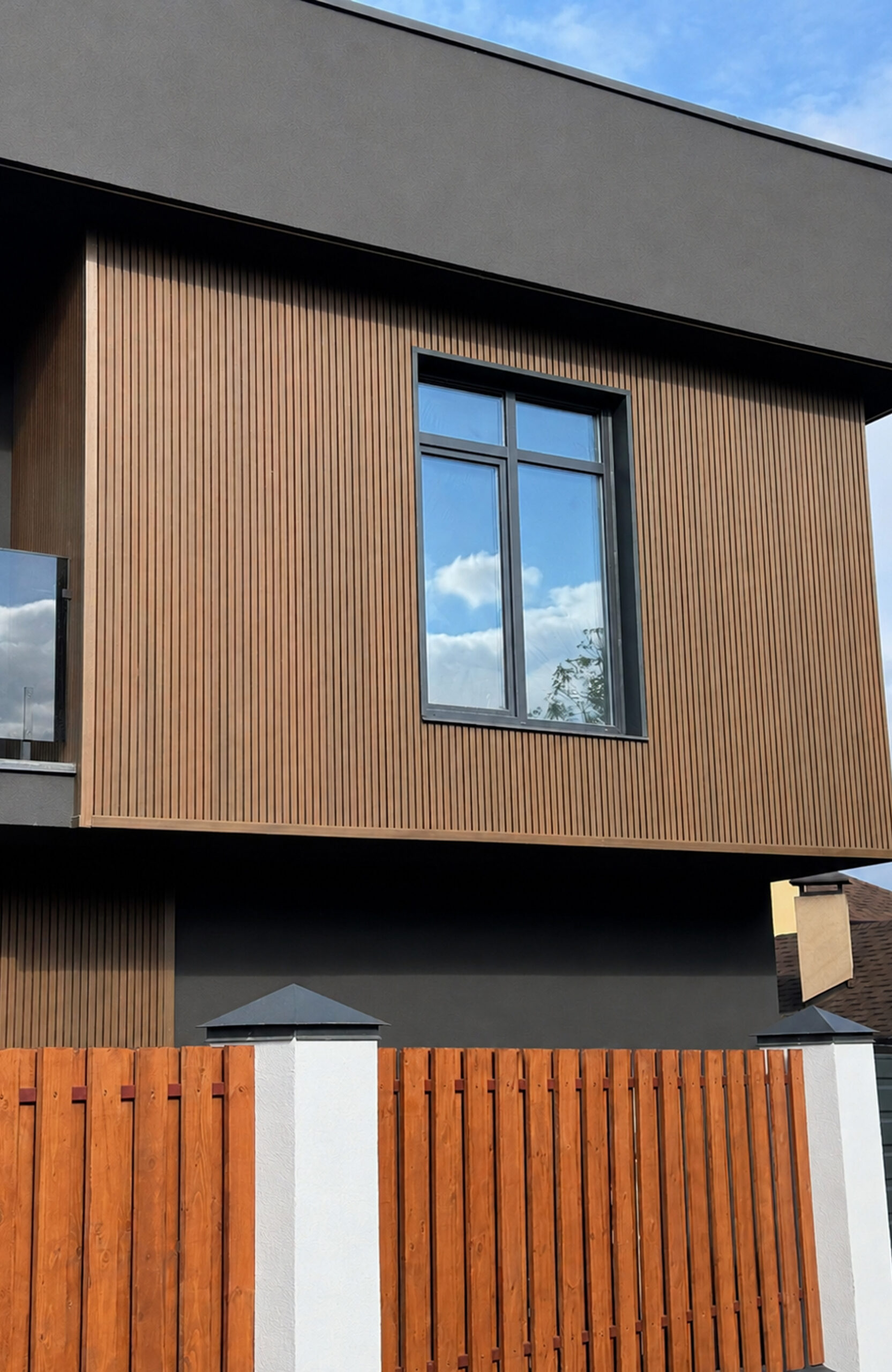 Modern house exterior with vertical wooden siding, a large window, and a gray overhanging roof/structure under a blue sky.