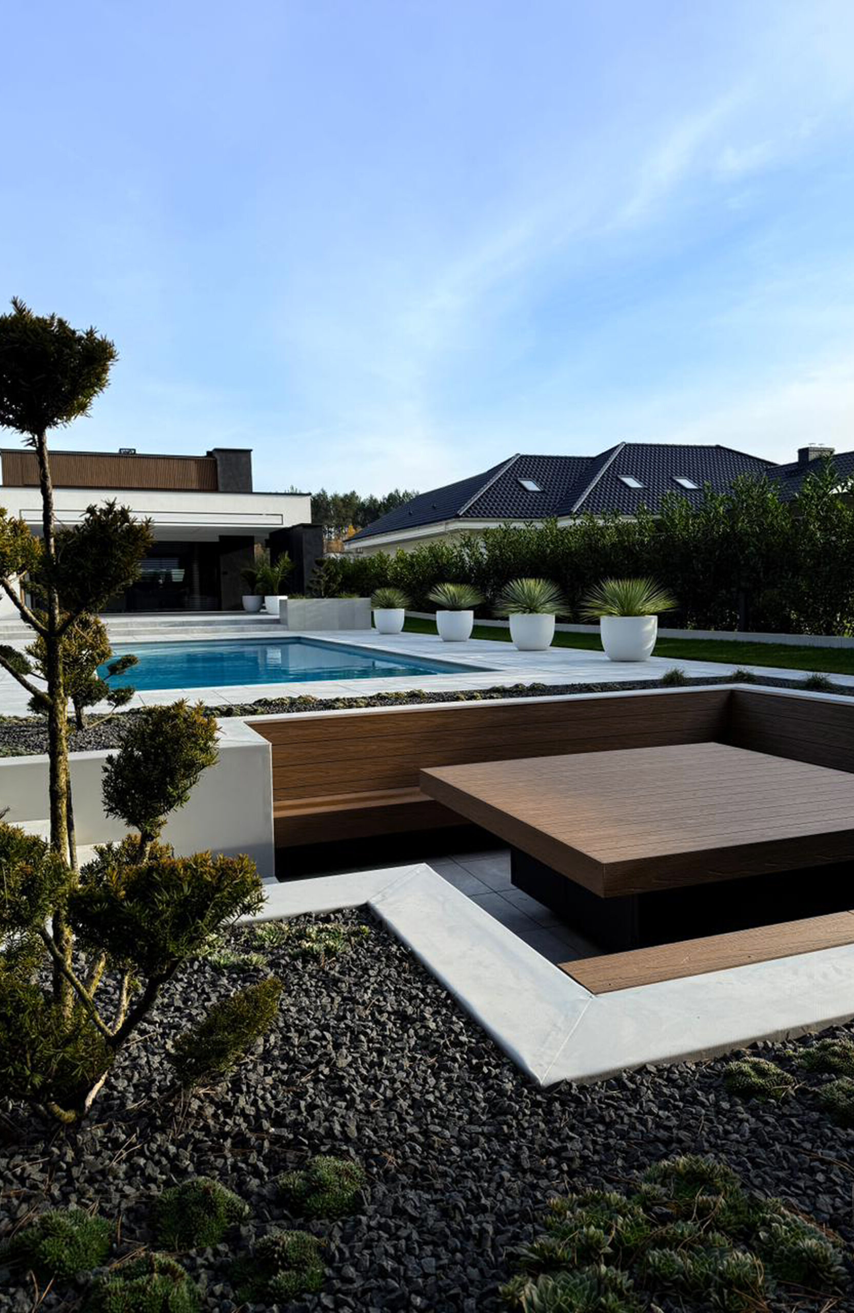 Modern backyard with a rectangular pool, white decking, and raised wooden seating areas surrounded by potted plants and gravel landscaping.