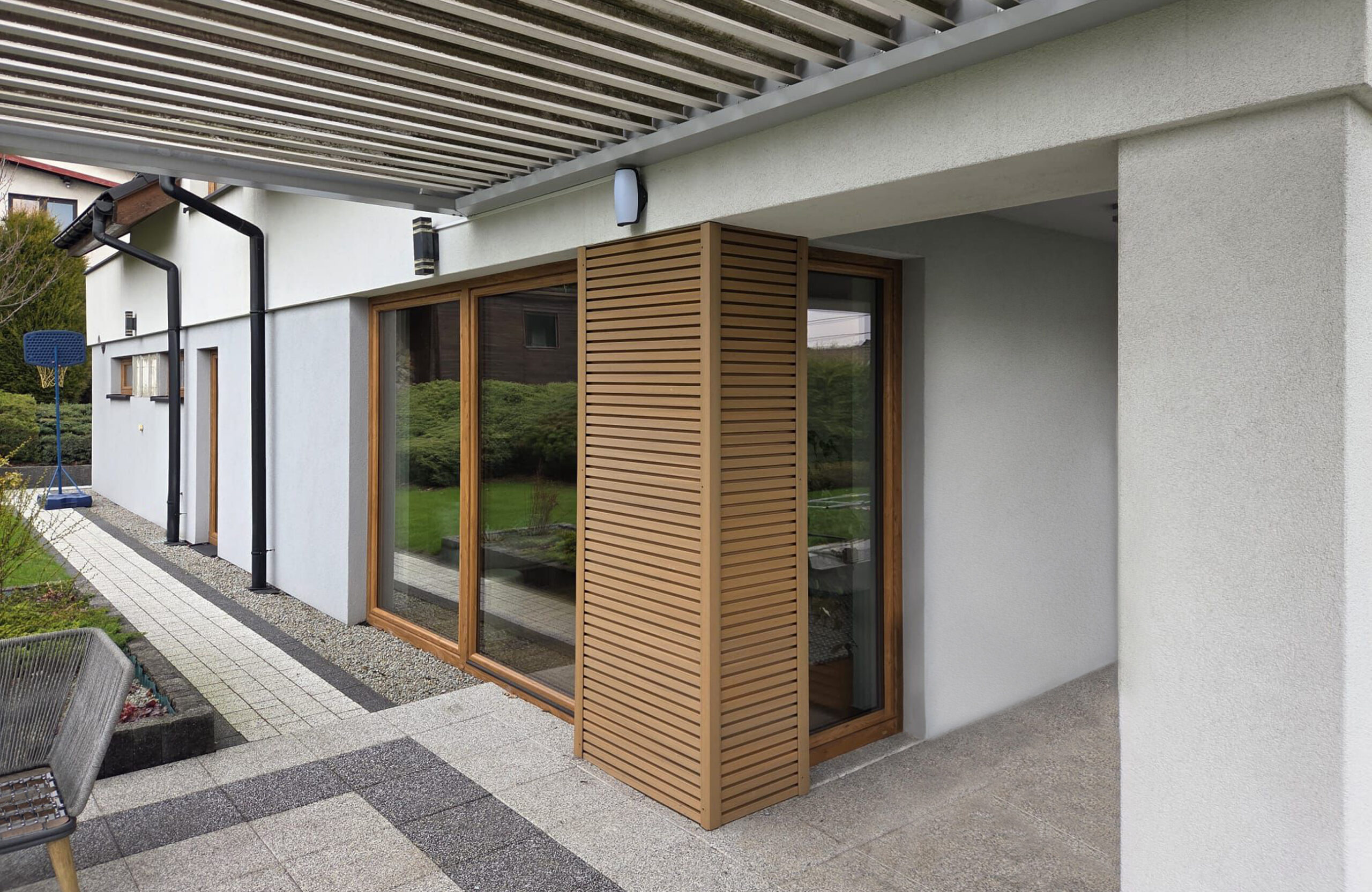 Modern house exterior with large glass doors, a wooden slatted privacy screen, and a metal pergola above; garden in background.