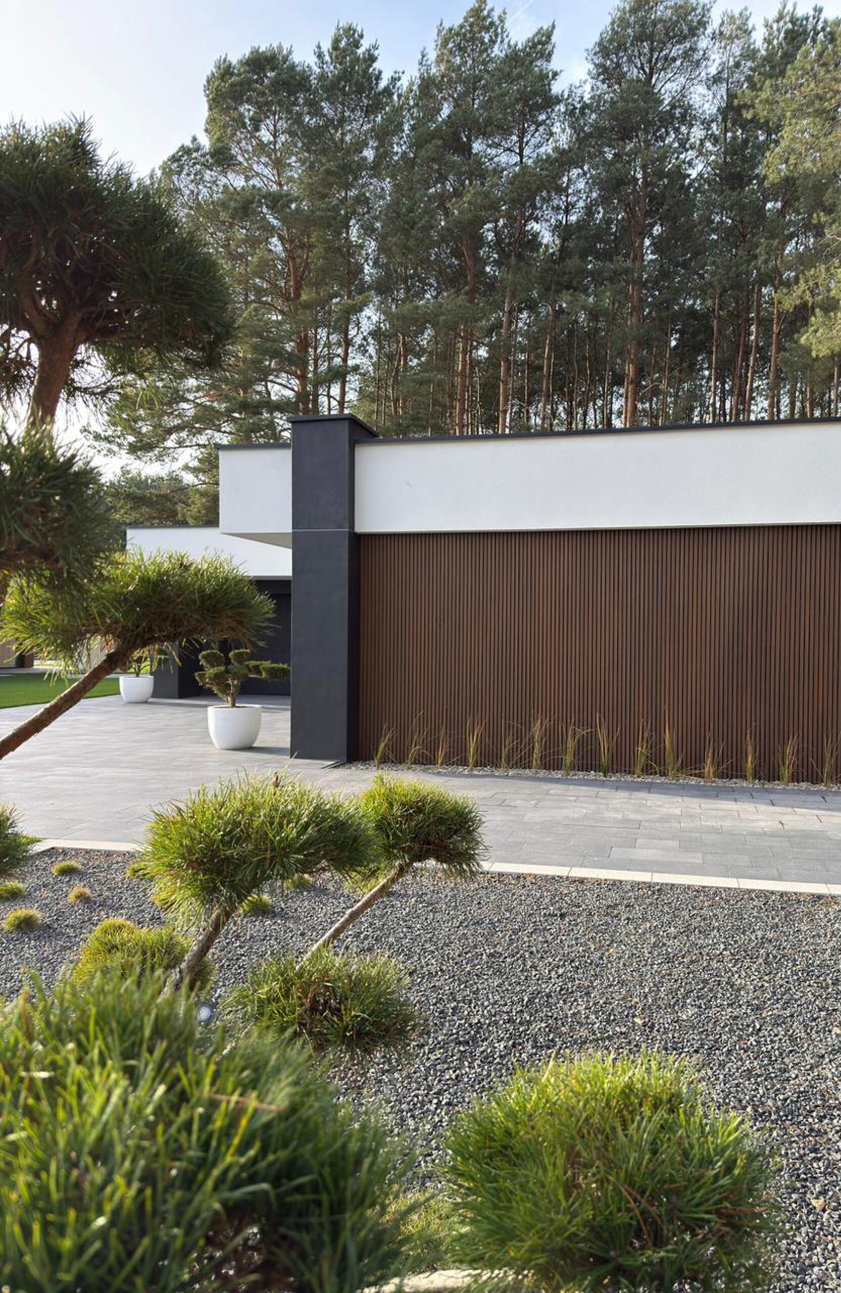 Modern minimalist house exterior with white walls, a dark vertical column, and a brown wooden slat fence, set behind a gravel and plant-filled yard with tall trees in the background