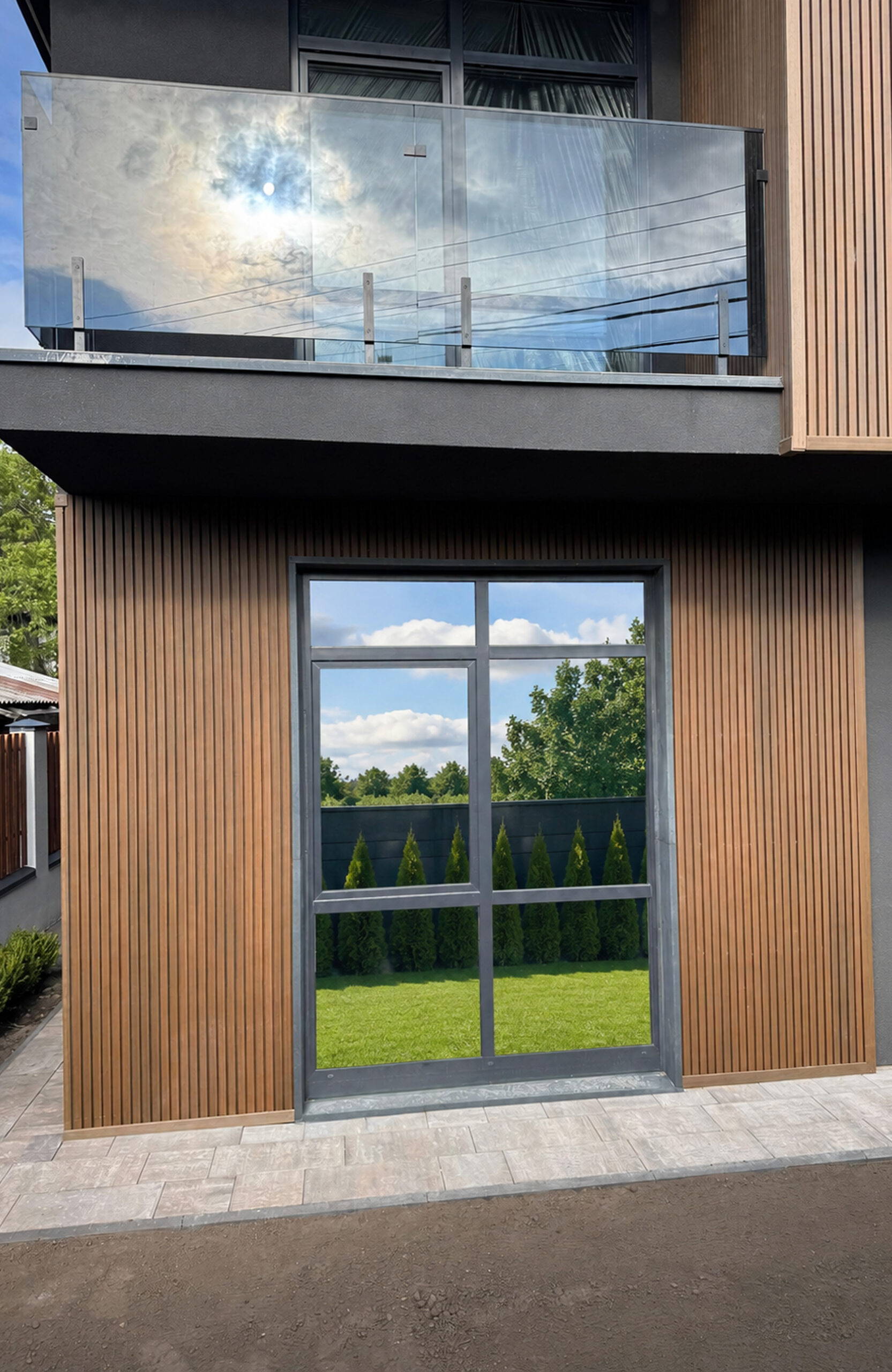 Modern house exterior featuring vertical wooden cladding, a glass balcony railing, and a large metal-framed window reflecting a blue sky. Behind the window is a green lawn and row of evergreen shrubs.