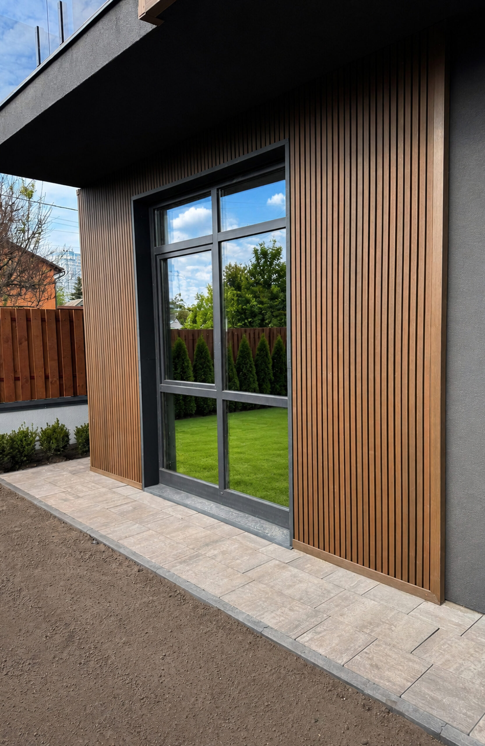 Modern exterior wall with vertical wooden slats around a large glass window; reflections of trees and sky in the glass.
