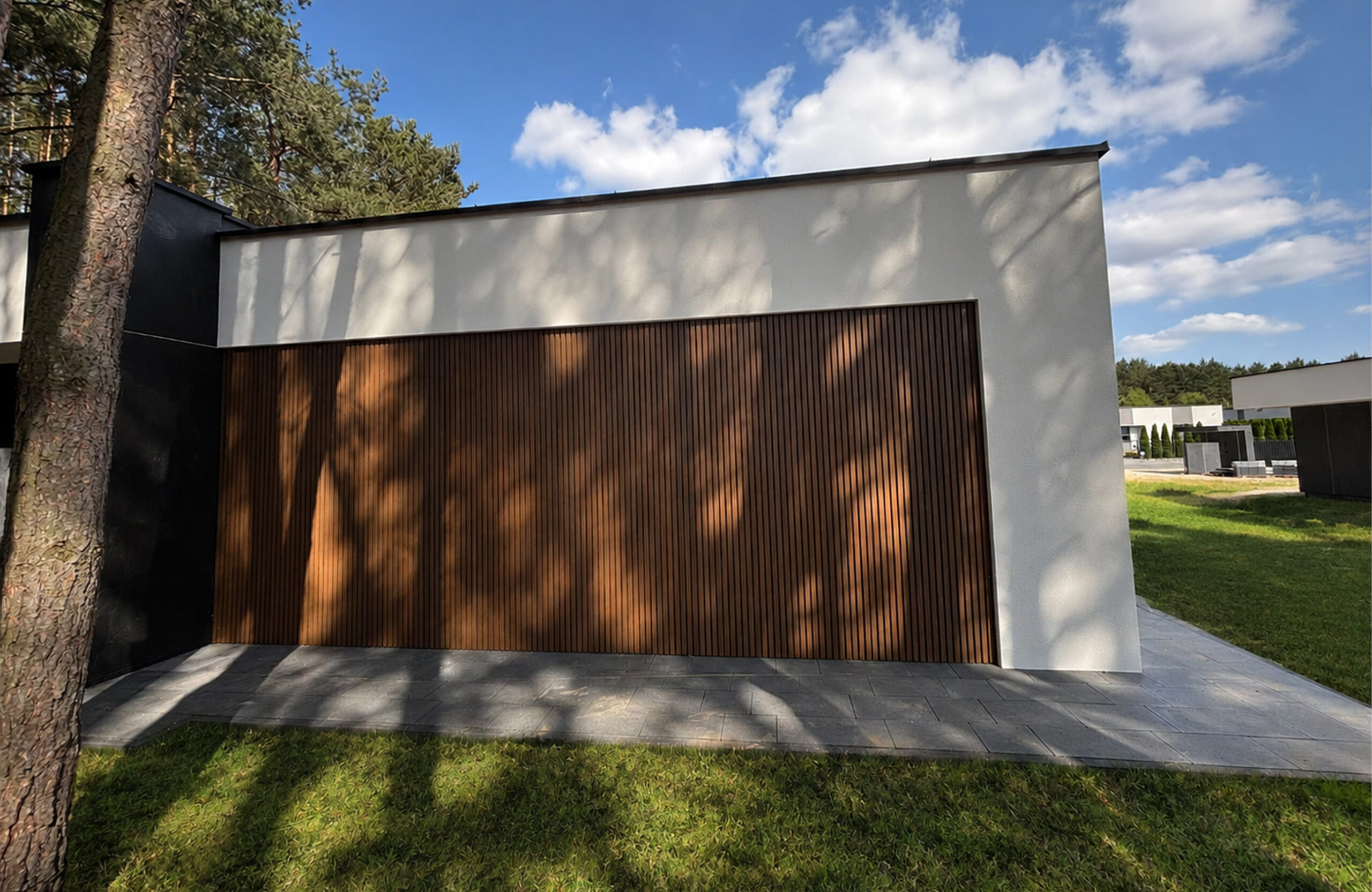 Modern white-walled house exterior with a vertical wooden slat garage door, shaded by a tall tree, under a blue sky with clouds.