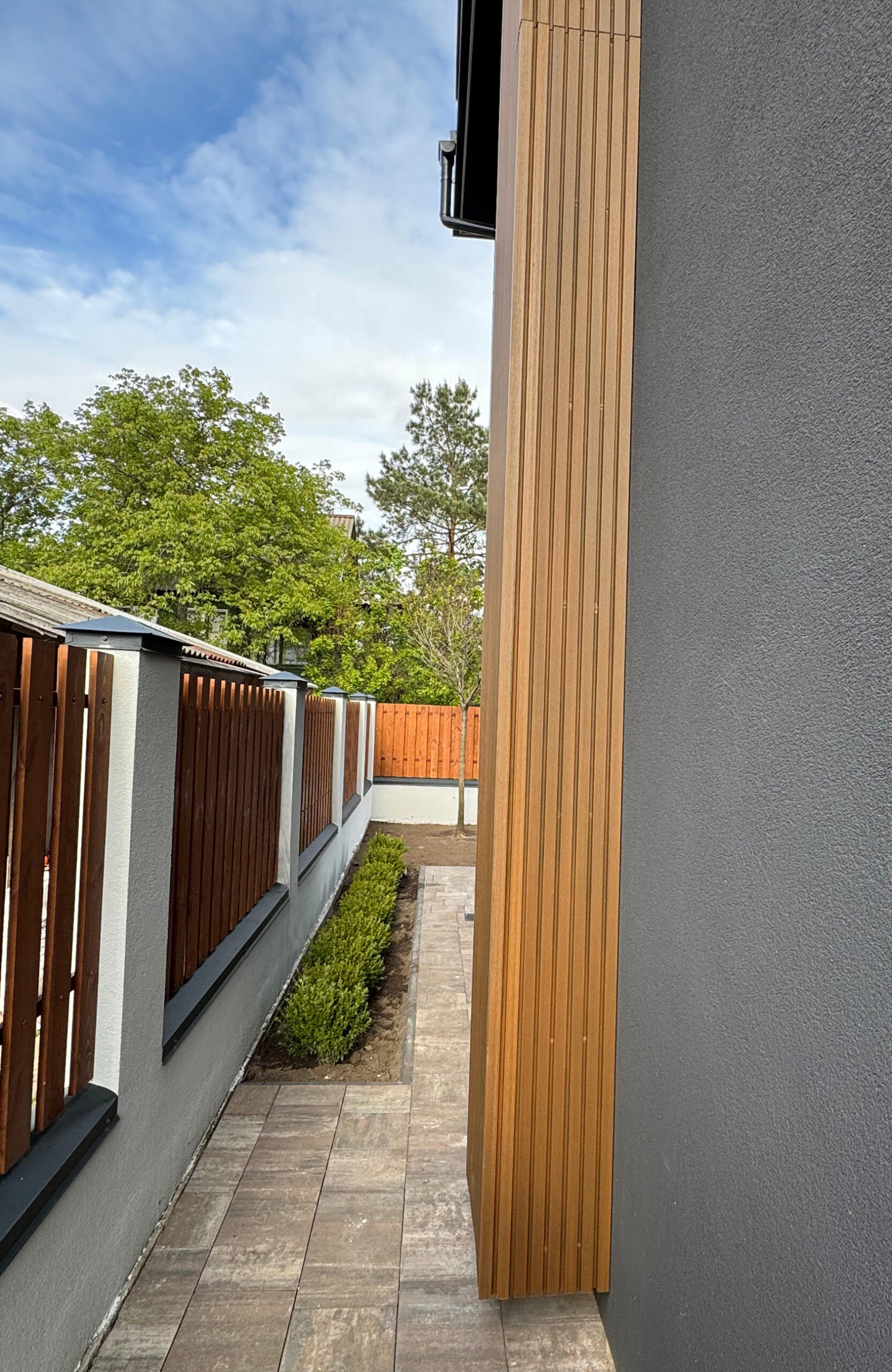 Narrow side walkway between a grey wall and vertical wooden slats, with a wooden fence and trees visible beyond.