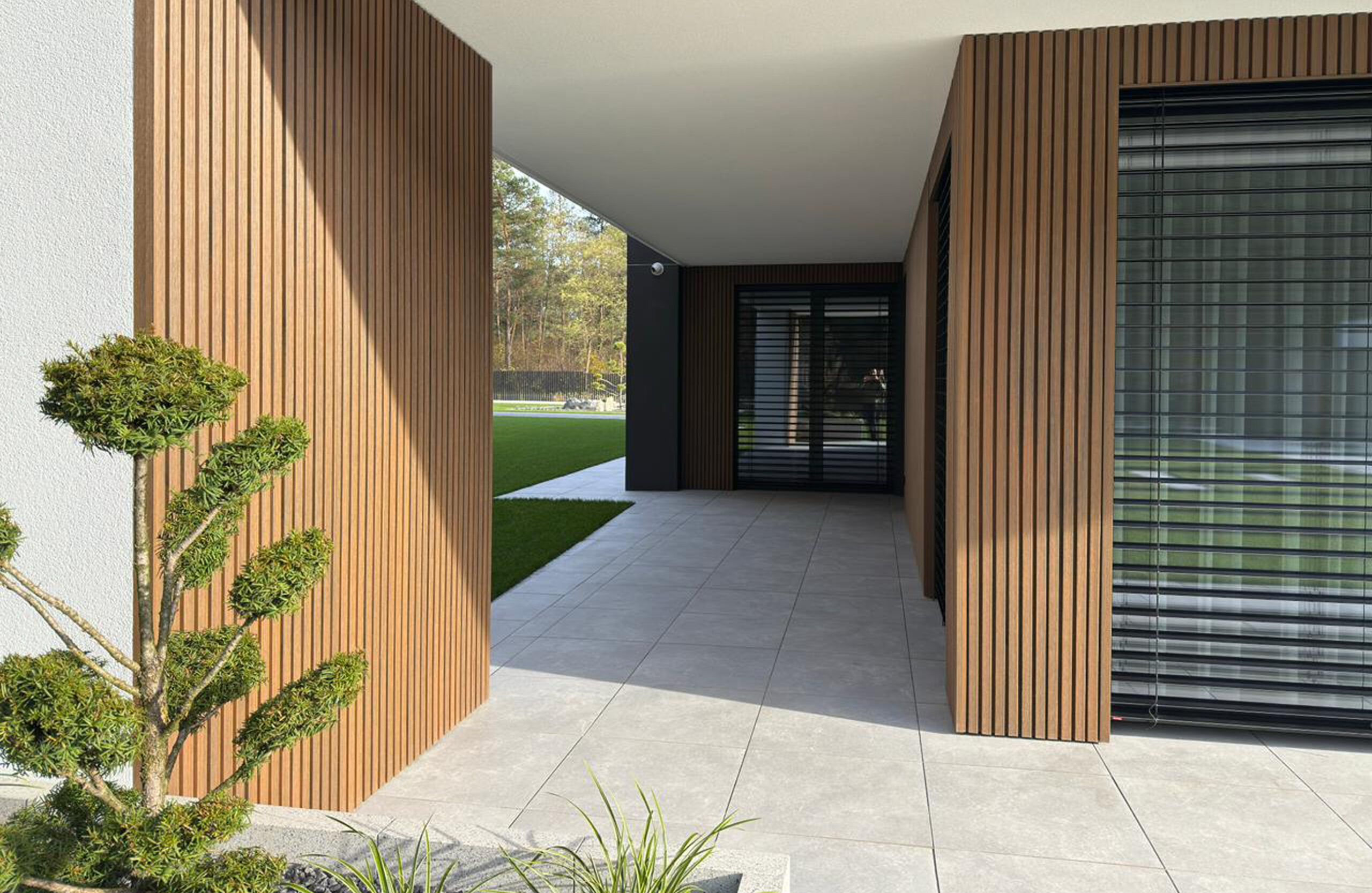 Modern house entrance featuring vertical wooden slats, a covered walkway, and a large window with blinds on the right.