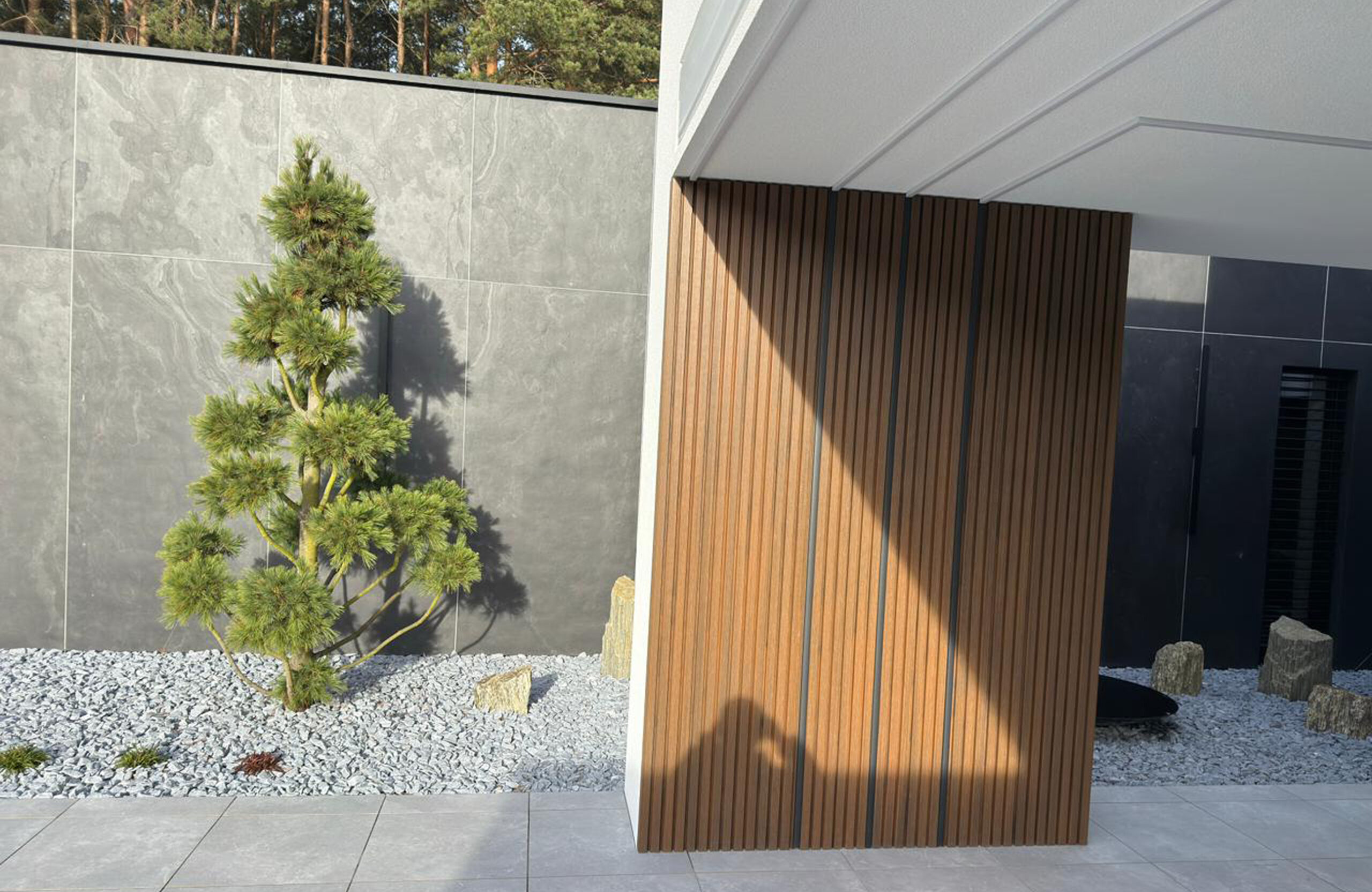 Modern outdoor space featuring a grey stone wall, vertical wooden slat screen, and a small pine tree in gravel ground.