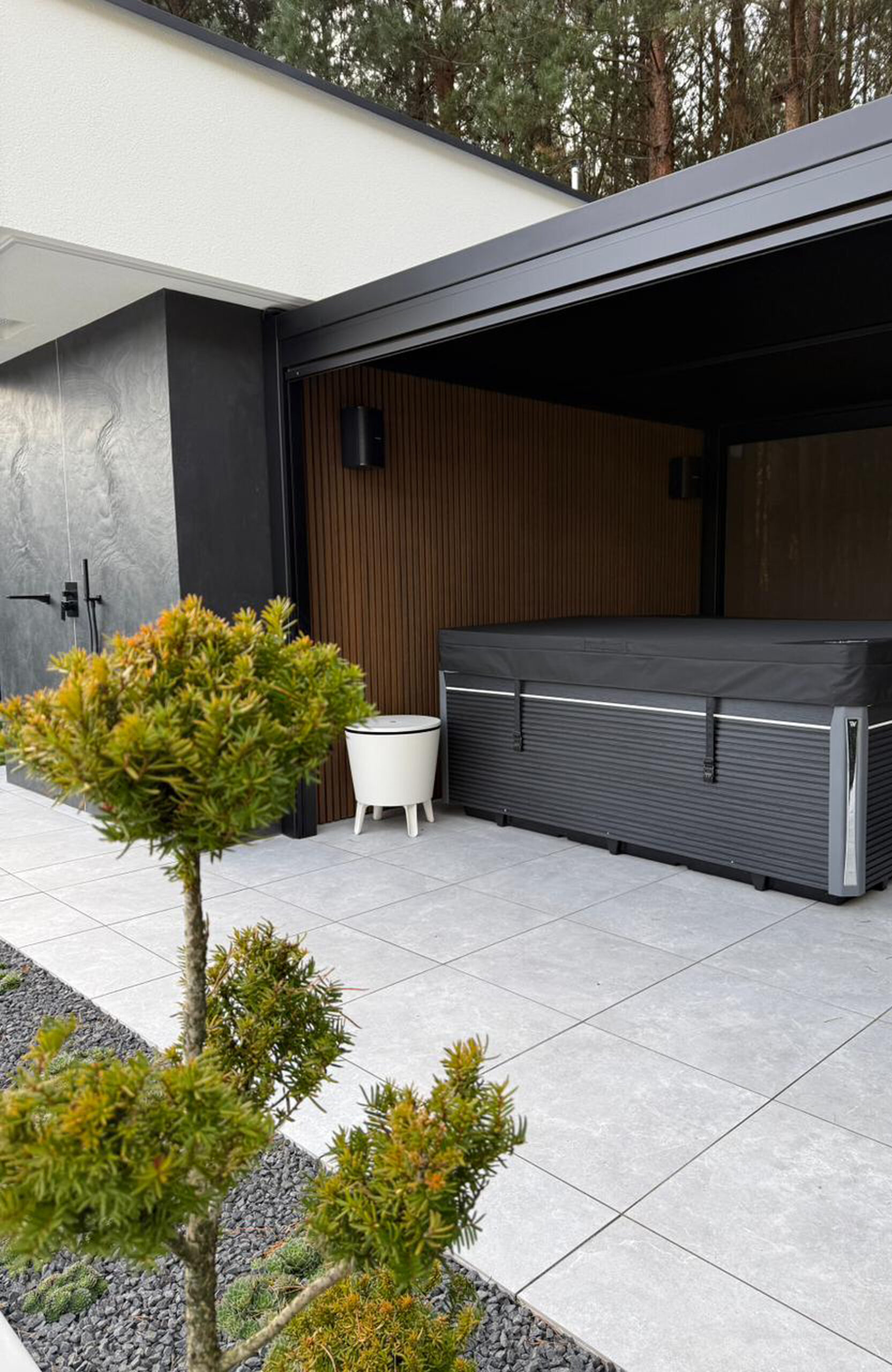 Modern outdoor patio with a large black grill cabinet along a wooden slat wall, gray tiled ground, and greenery in the foreground.",