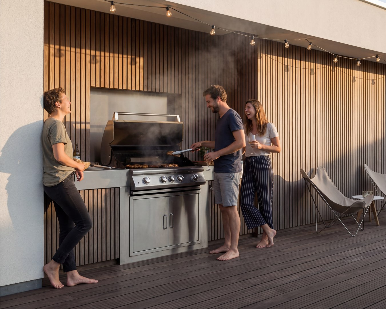 Terrace Near a Barbecue Area: Which Decking Should You Choose and What Should You Avoid? Outdoor kitchen with built-in stainless steel gas grill and friends enjoying BBQ on wooden terrace