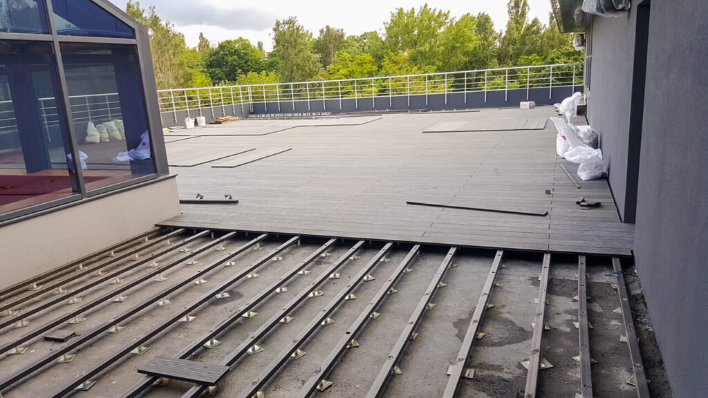 Rooftop deck under construction with exposed metal joists and partially laid wooden planks; glass-walled room on the left and bags of materials along the wall.