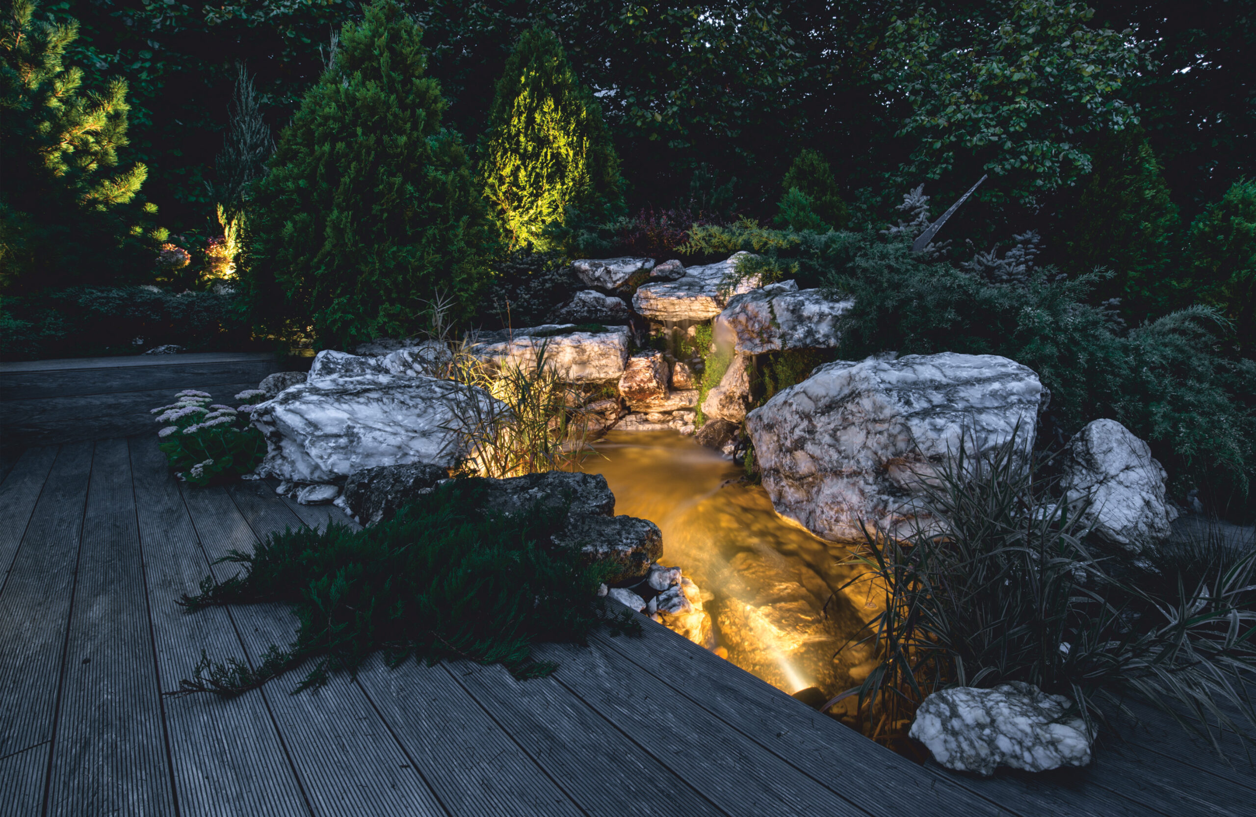 Landscaped outdoor area with BRUGGAN Multicolor grey composite decking, lush plants, rocks and water features