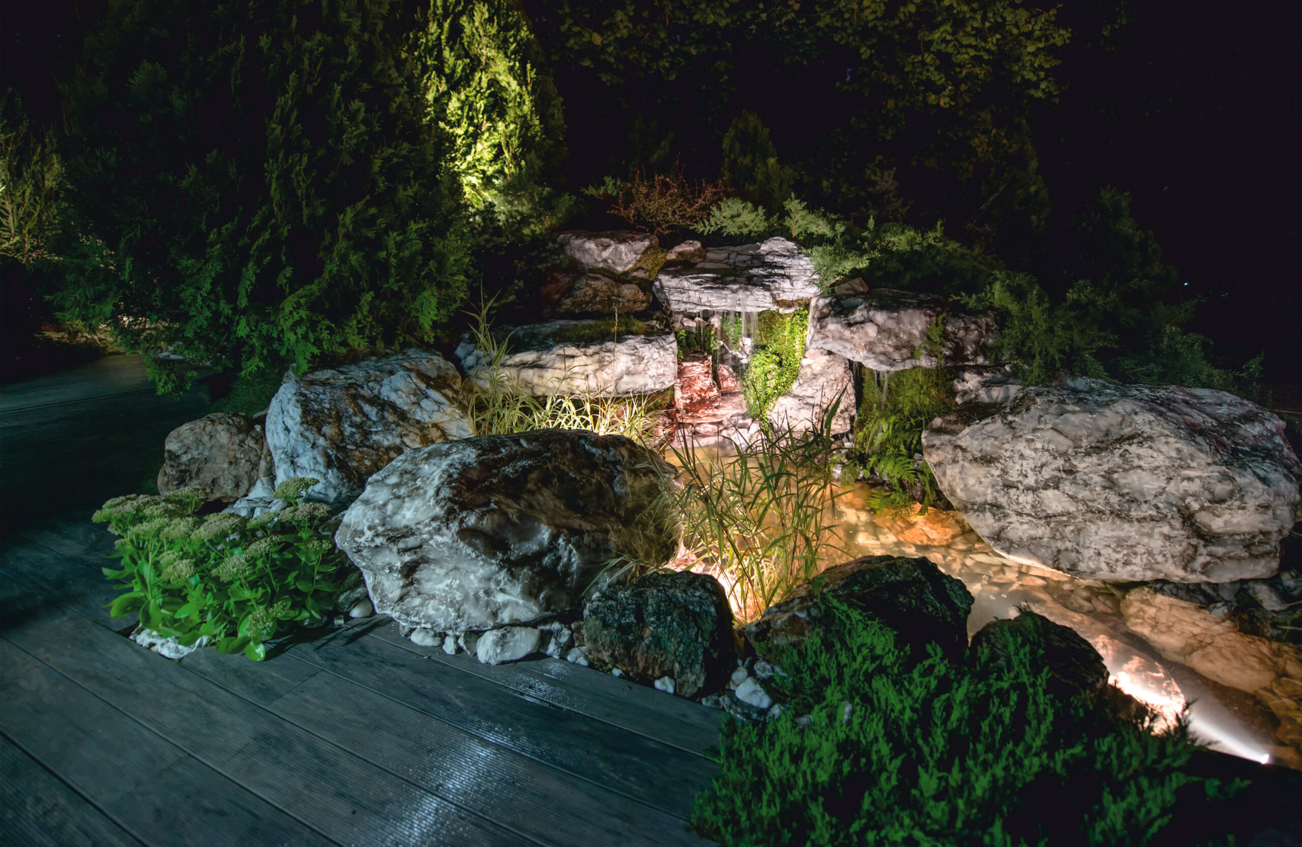Landscaped outdoor area with BRUGGAN Multicolor grey composite decking, lush plants, rocks and water features