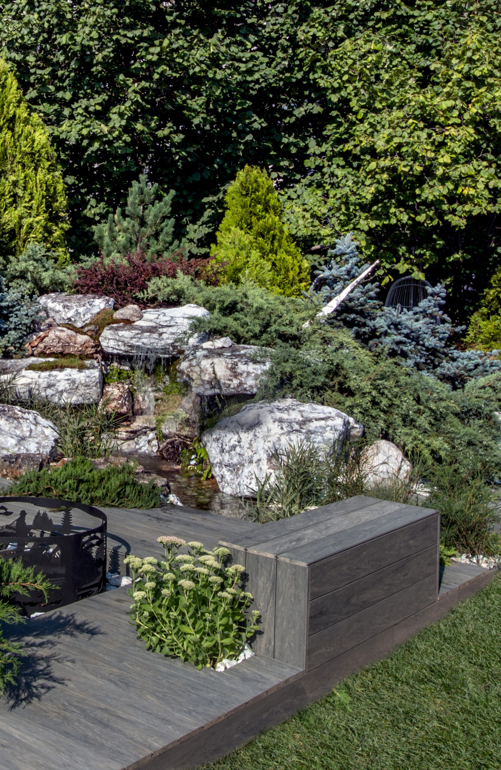 Landscaped outdoor area with BRUGGAN Multicolor grey composite decking, lush plants, rocks and water features
