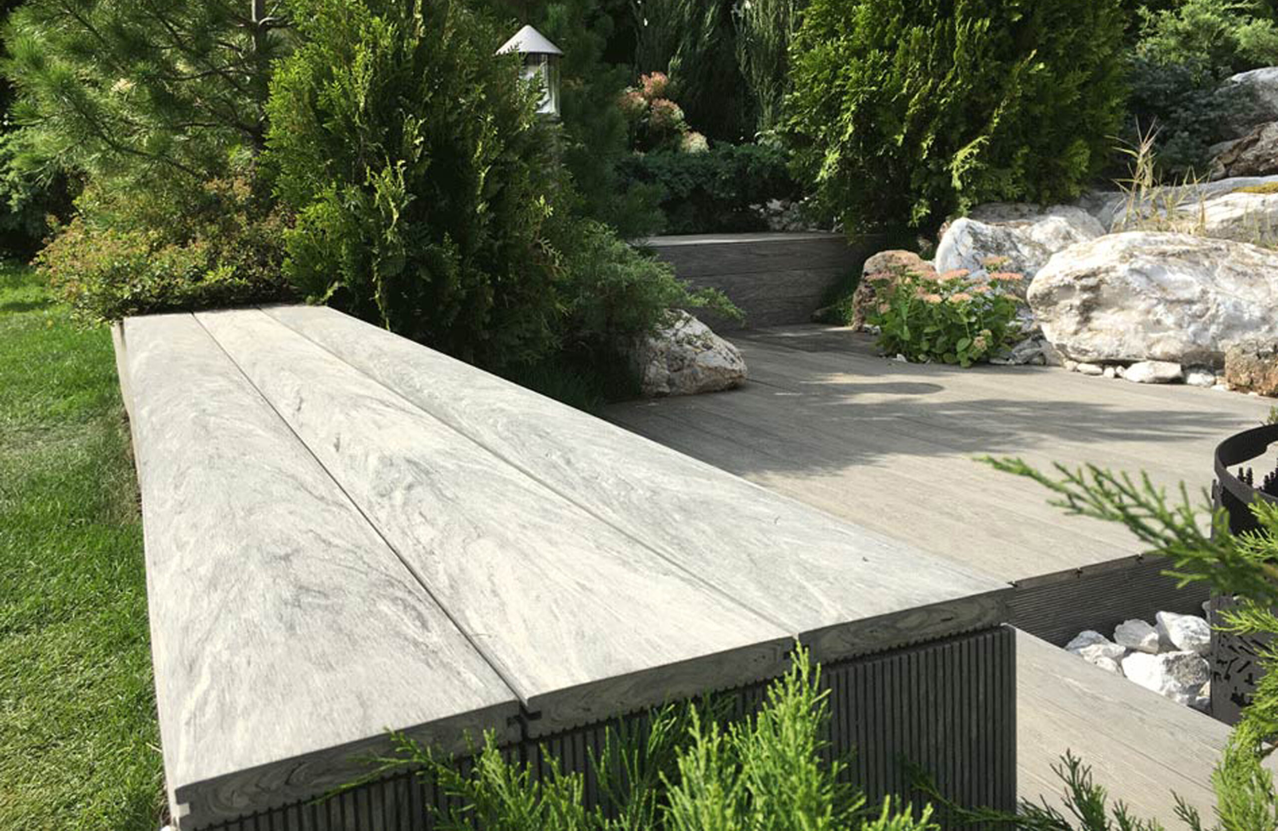 Landscaped outdoor area with BRUGGAN Multicolor grey composite decking, lush plants, rocks and water features