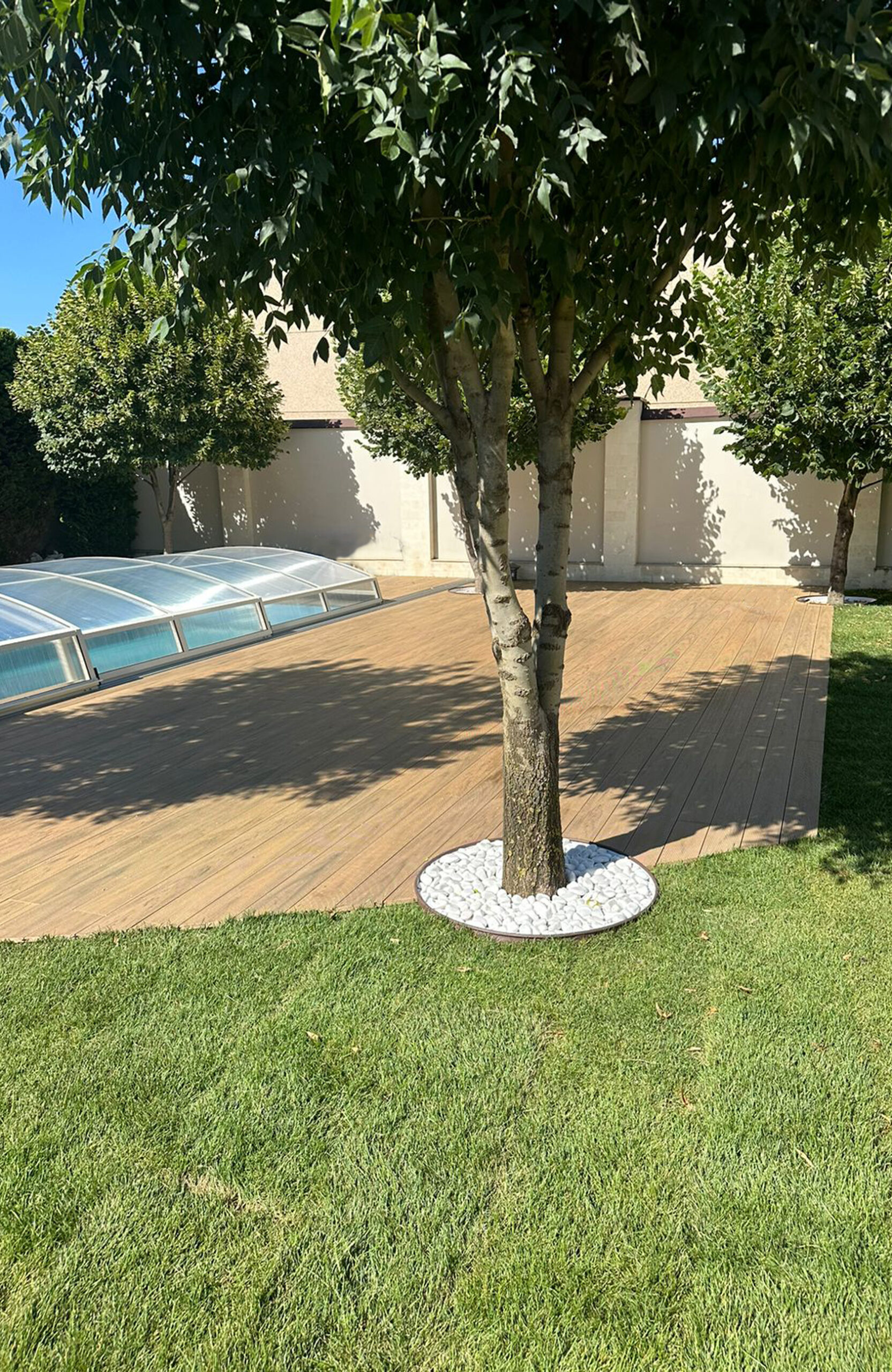 Sand-colored BRUGGAN Multicolor composite decking surrounding a garden pool with trees and lawn