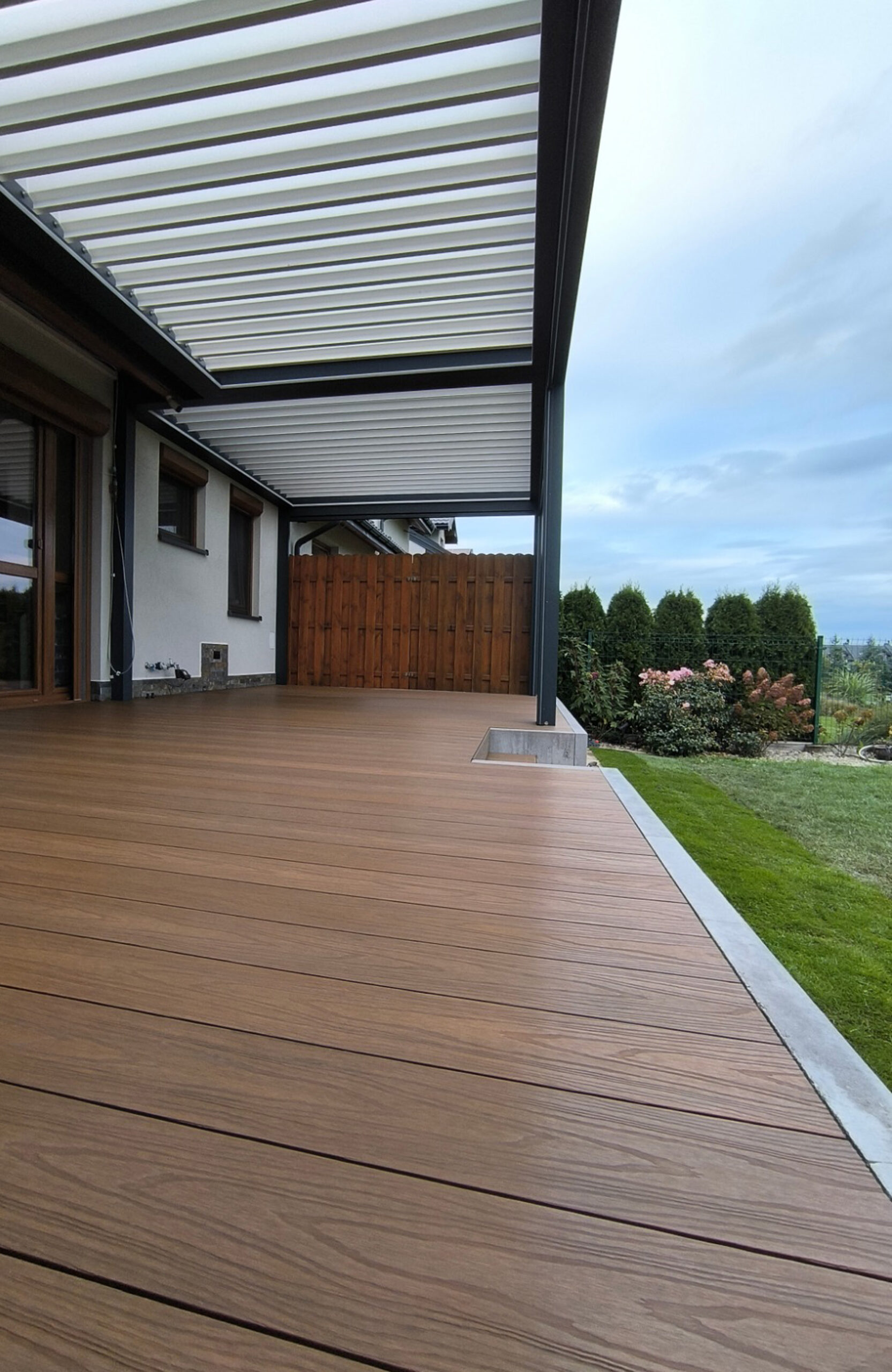 Spacious backyard terrace featuring natural wood-grain composite decking, a dark grey louvered pergola, and a wooden privacy fence next to a green lawn