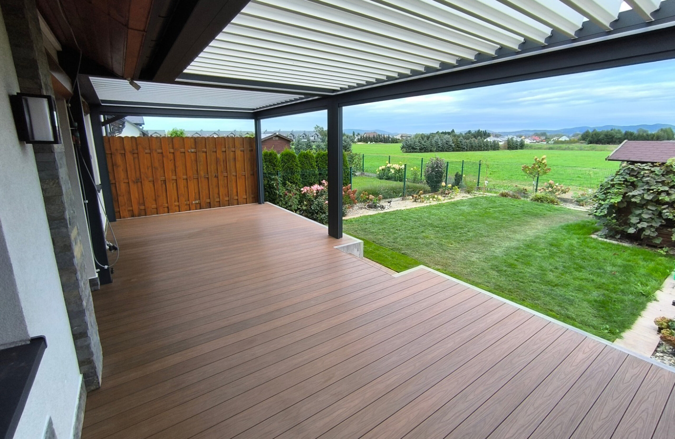 Spacious backyard terrace featuring natural wood-grain composite decking, a dark grey louvered pergola, and a wooden privacy fence next to a green lawn
