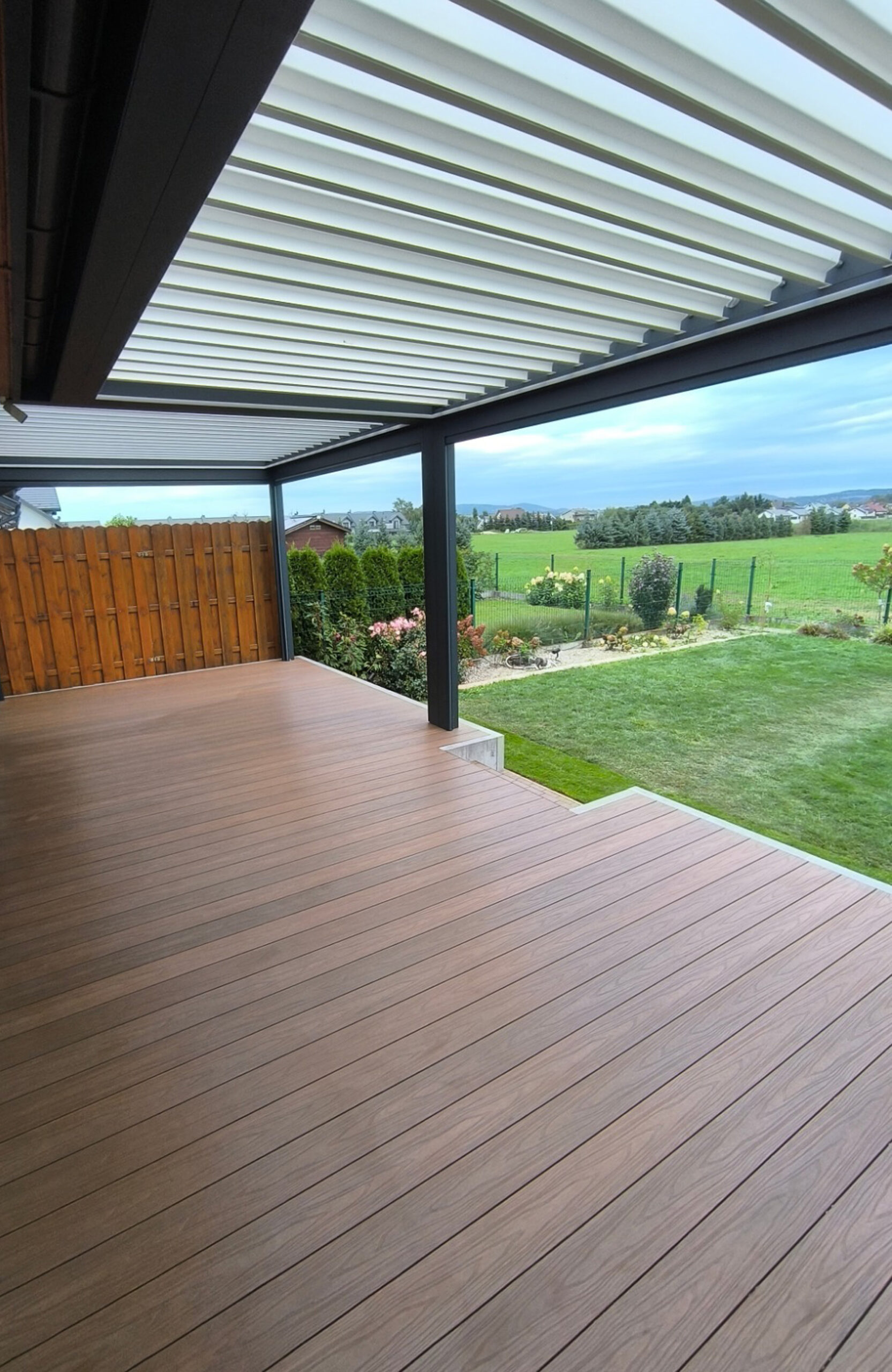 Spacious backyard terrace featuring natural wood-grain composite decking, a dark grey louvered pergola, and a wooden privacy fence next to a green lawn