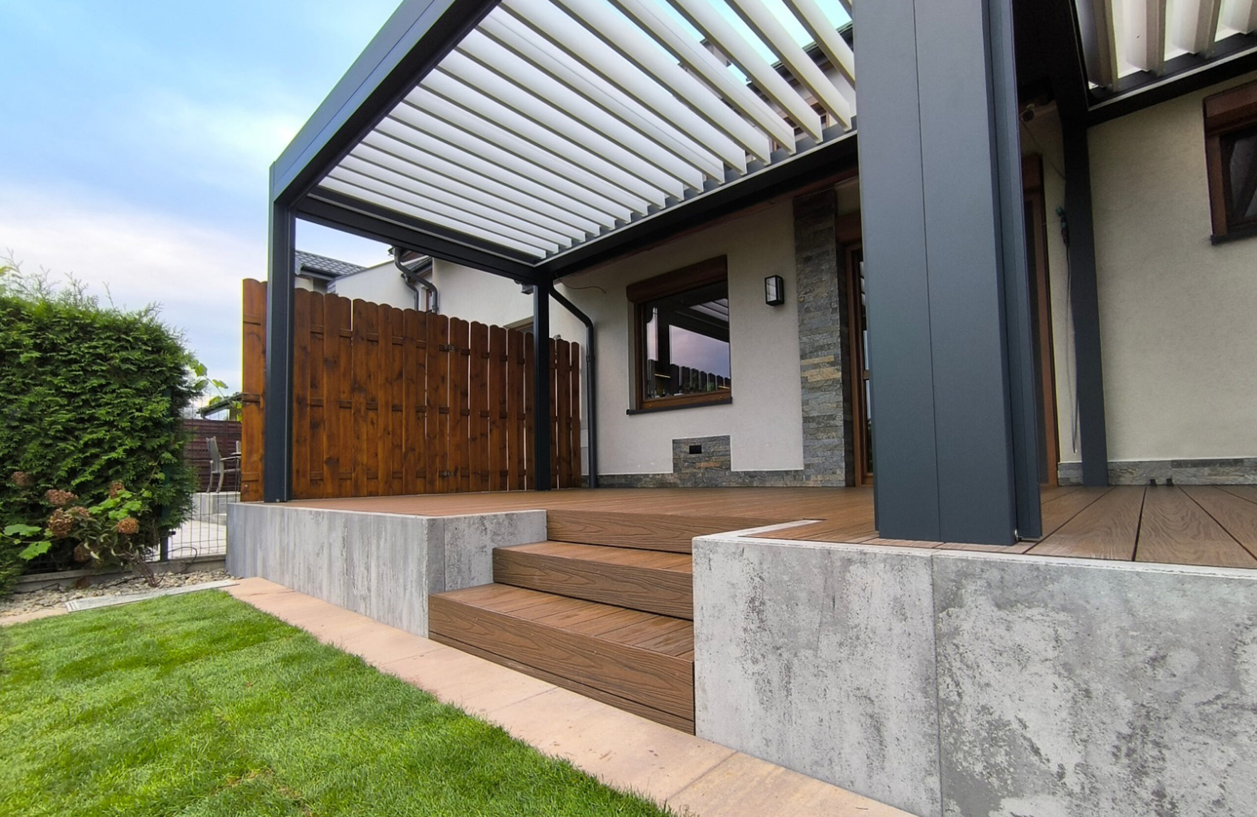 Modern outdoor terrace entry featuring composite wood steps, concrete retaining walls, and a black aluminum pergola with adjustable roof louvers