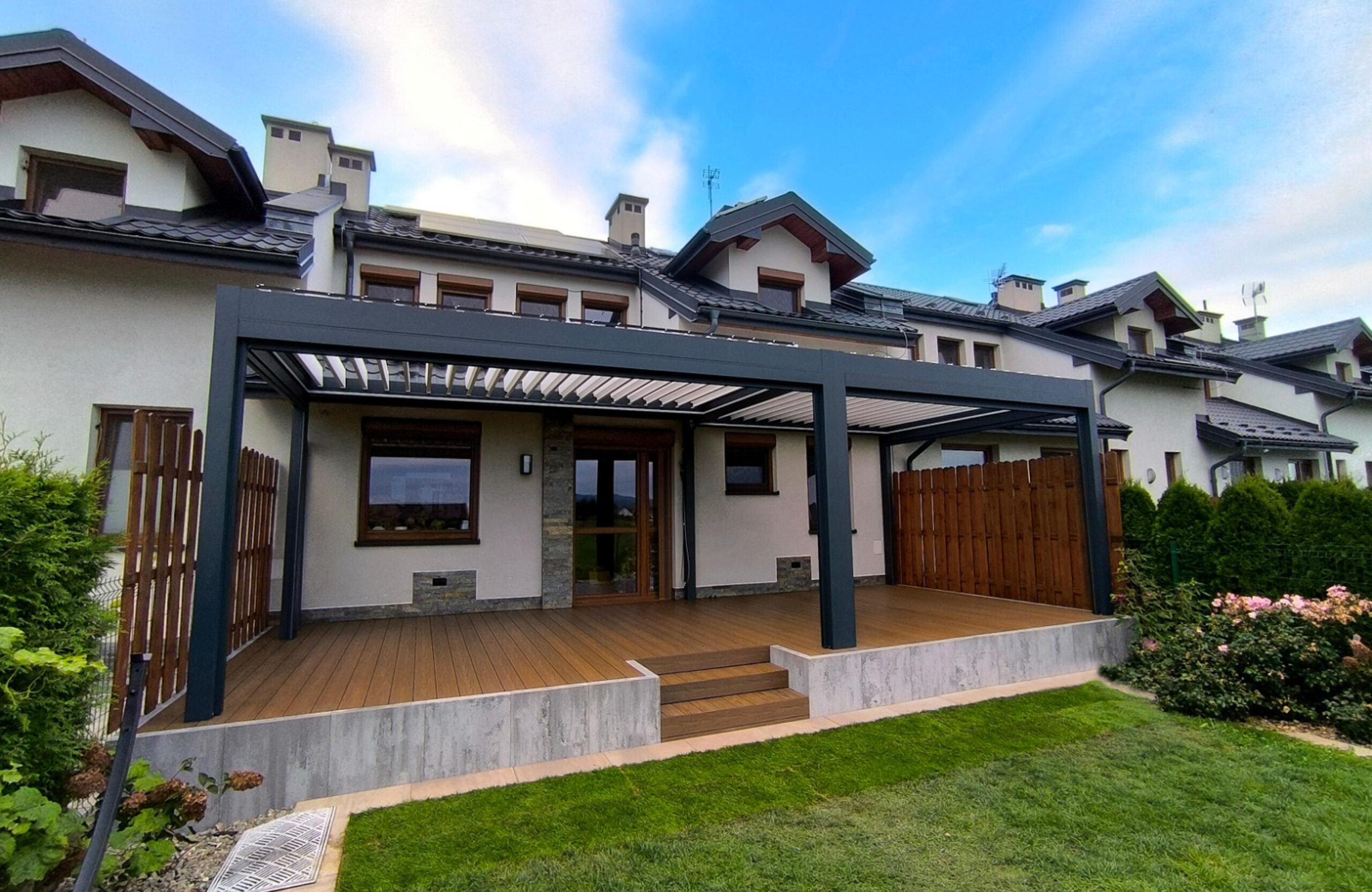 Exterior view of a townhouse patio with warm brown composite decking, integrated composite steps, concrete base foundation, and a full-length black louvered pergola roof
