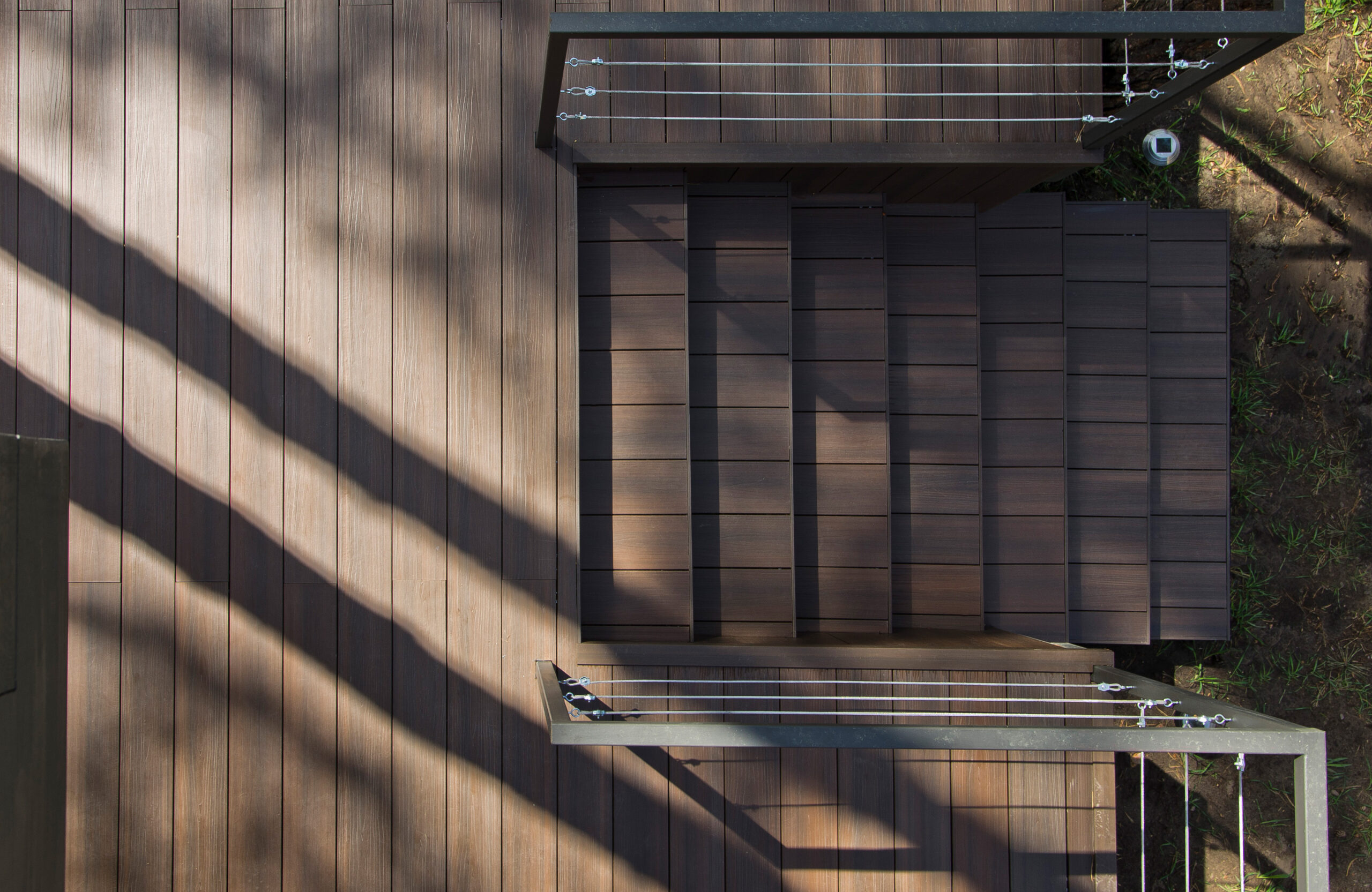 Top-down view of a modern outdoor composite deck in a deep walnut color, with stairs, a metal cable railing, and distinct diagonal shadows from overhead structures