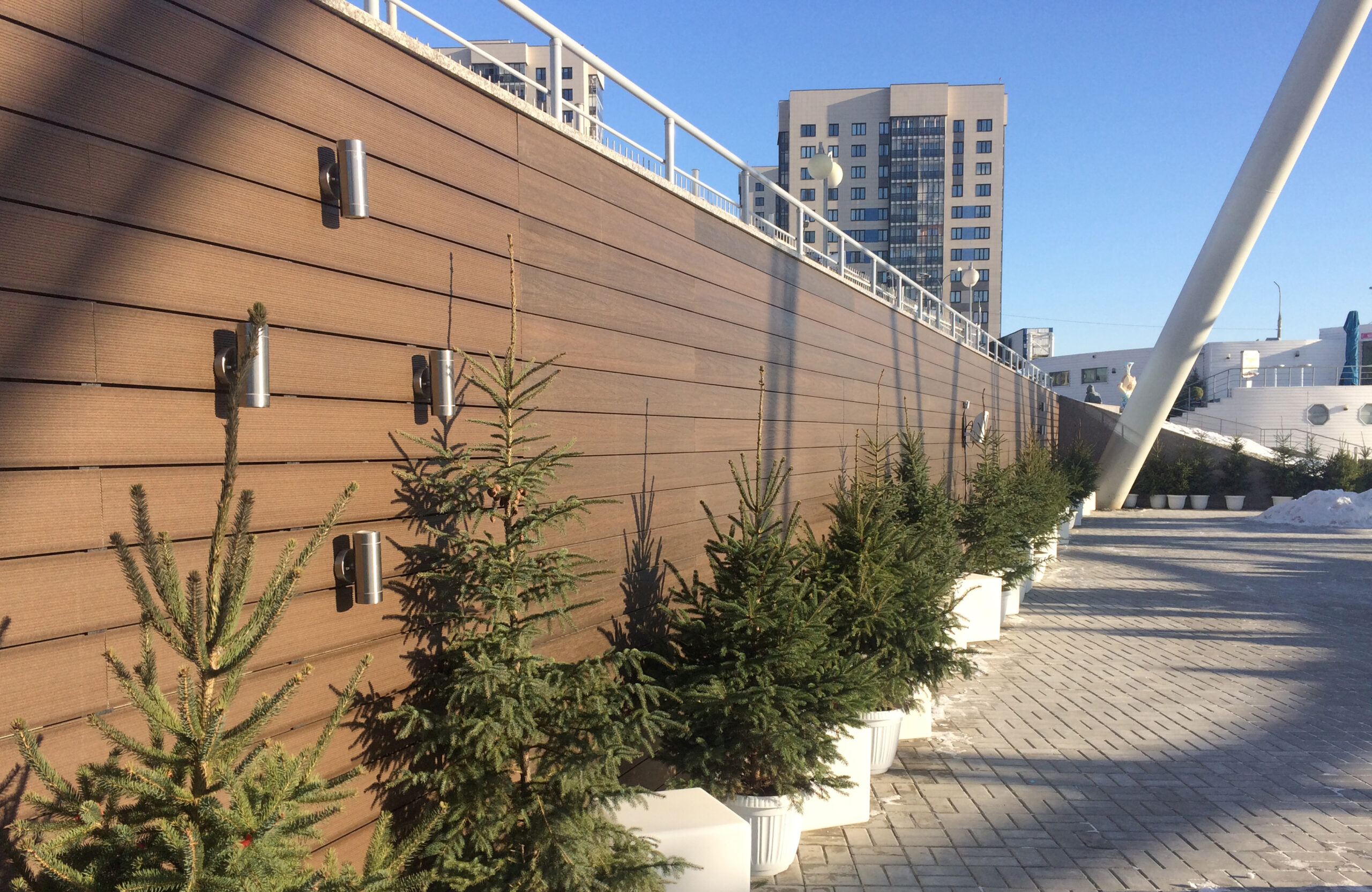 Brown composite wall cladding with outdoor lighting and evergreen trees in a modern urban setting