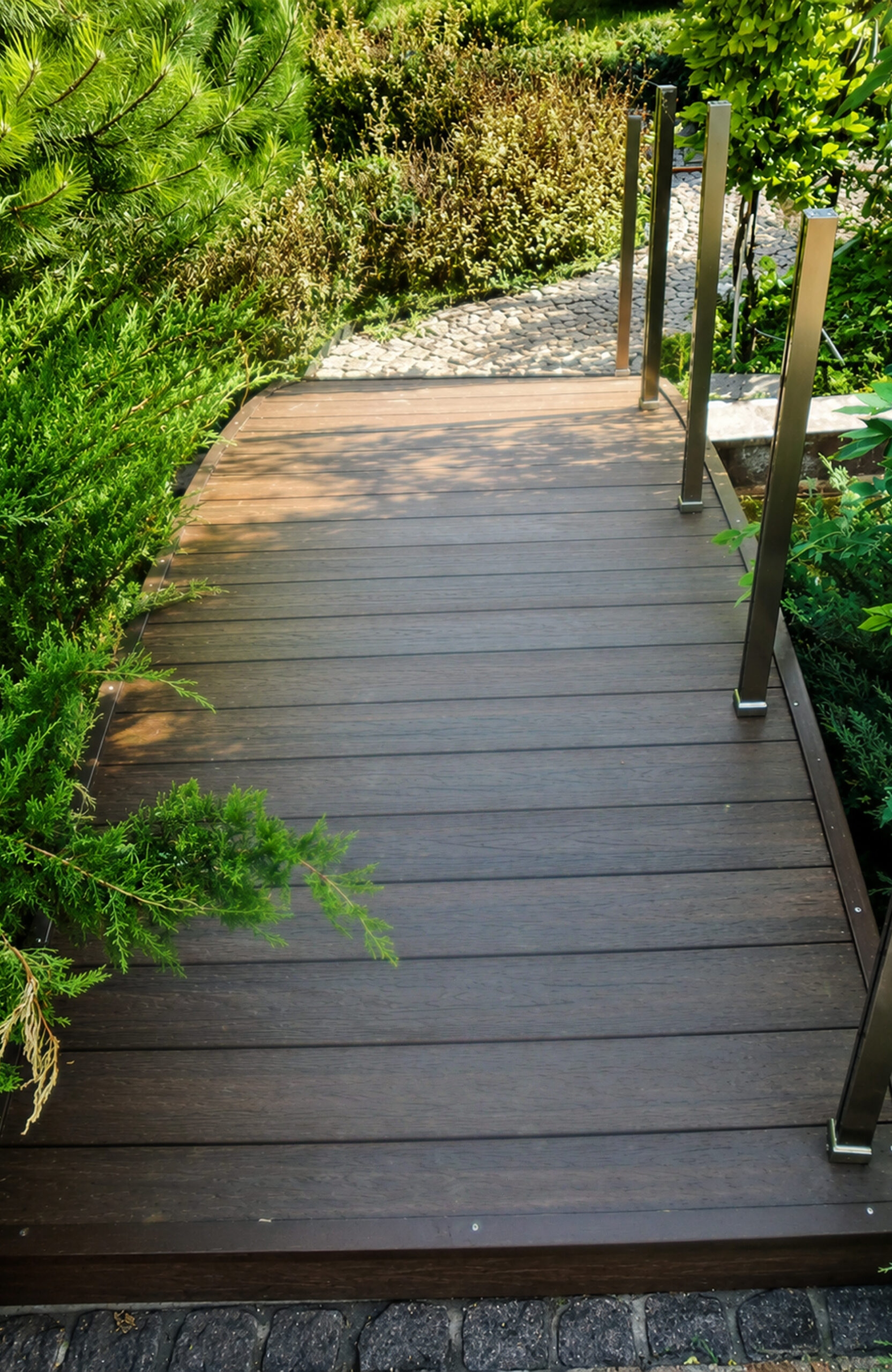 A long-angle perspective view of a garden bridge or walkway with rich walnut composite decking boards, framed by diverse green bushes and evergreens, with a stone path visible beyond