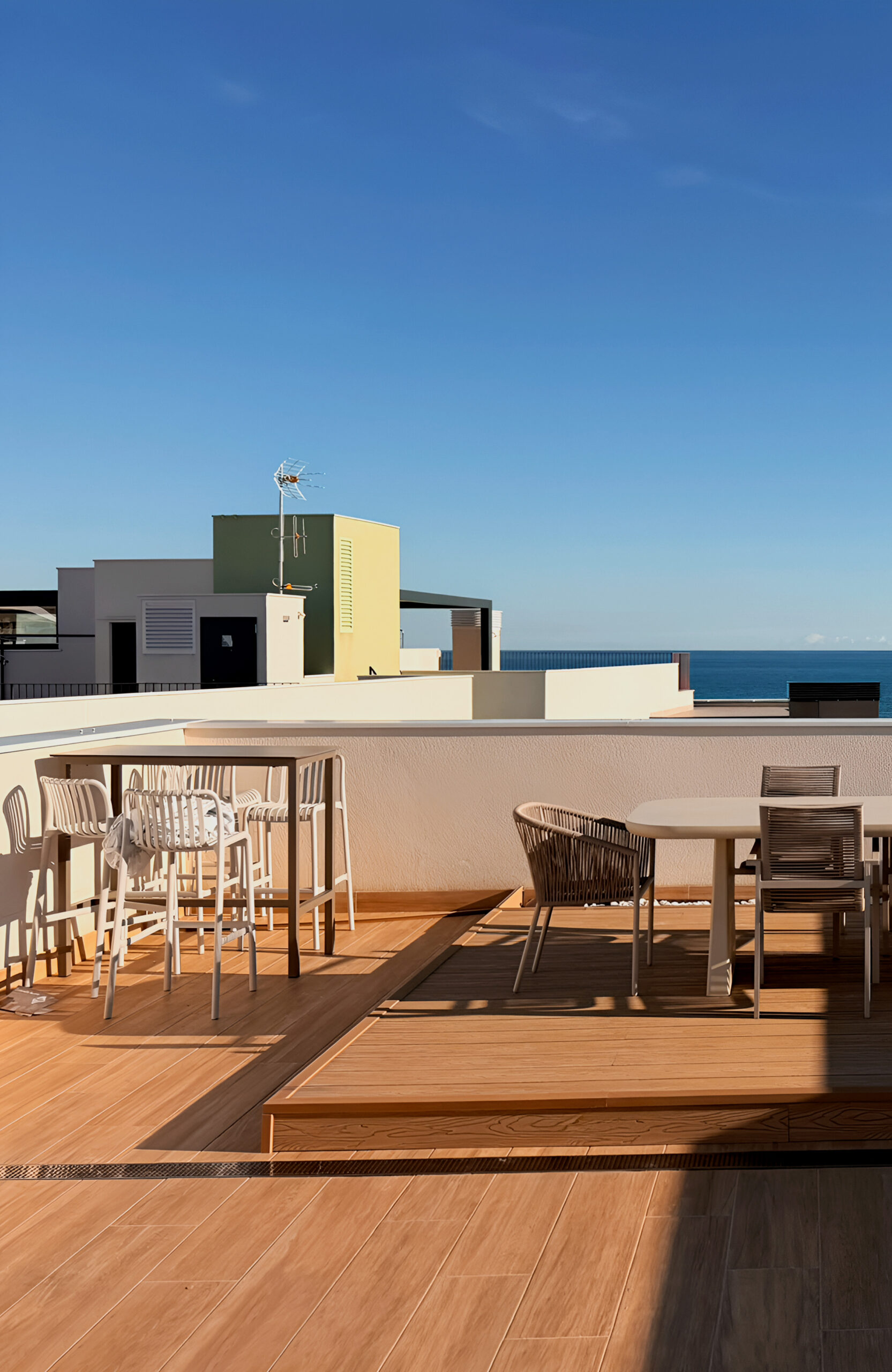 A modern rooftop terrace with outdoor dining furniture under a clear blue sky overlooking the sea